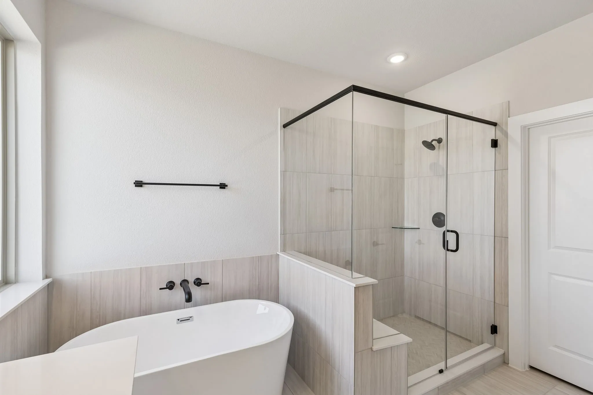 Freestanding soaking tub with wall-mounted matte black faucet, glass-enclosed shower with matte black fixtures, and light-toned tile flooring