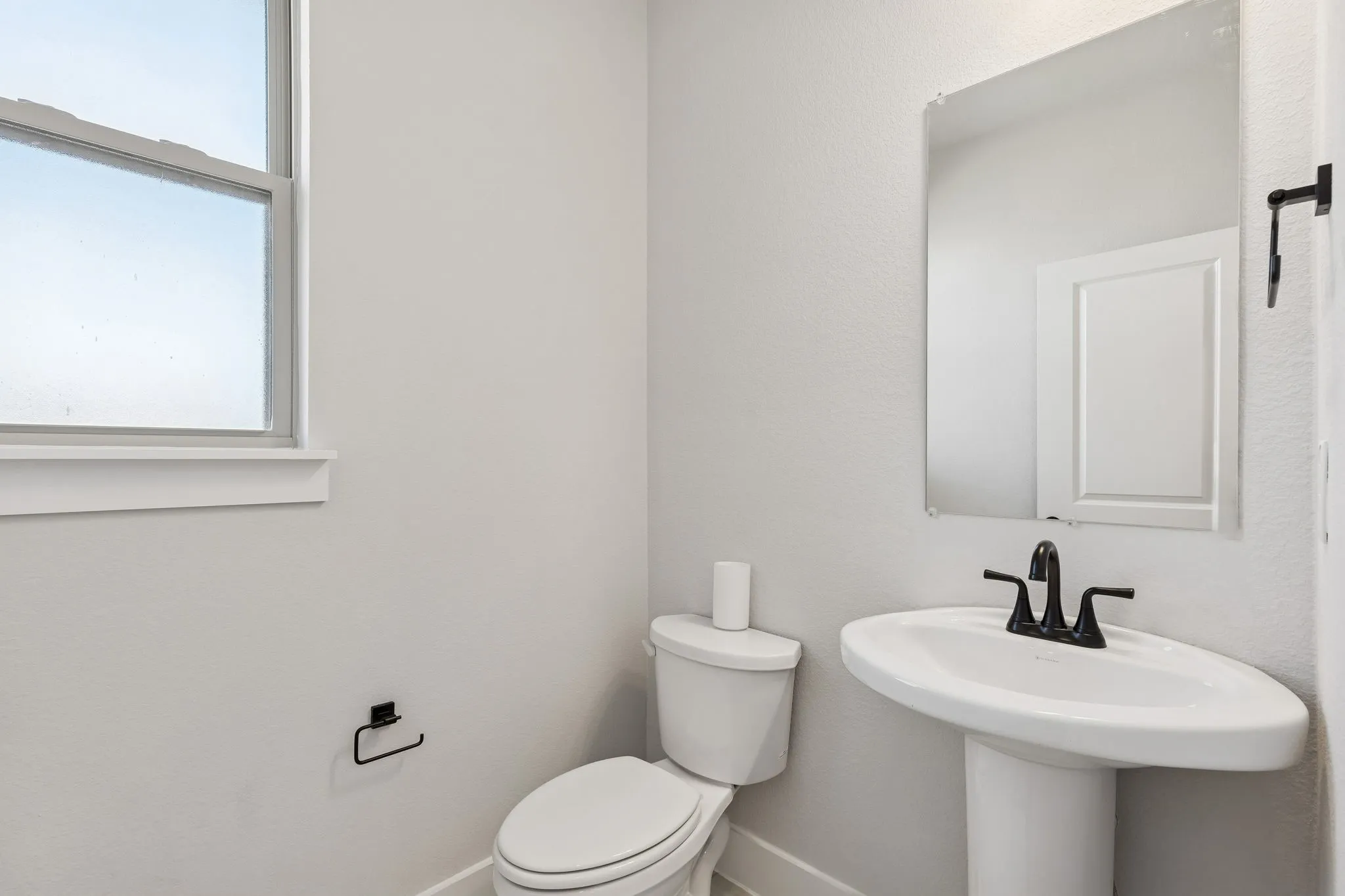 Pedestal sink with a matte black faucet, wall-mounted mirror, and a window with white trim