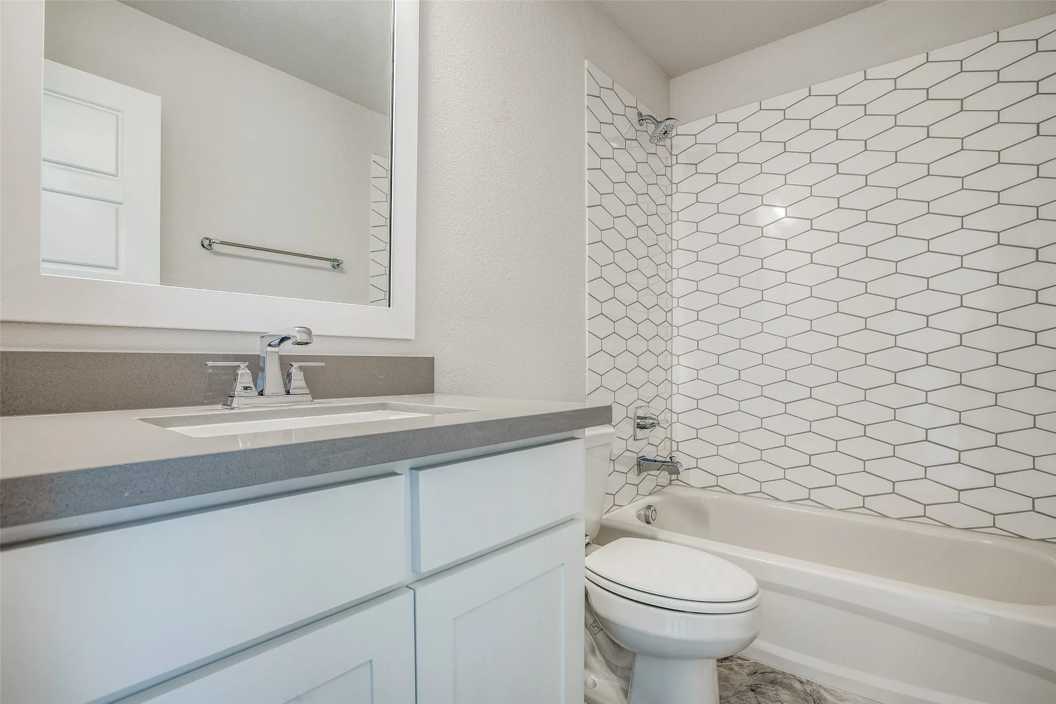 White shaker cabinetry, grey countertop, undermount sink with chrome faucet, hexagon-patterned tub surround, and a white bathtub