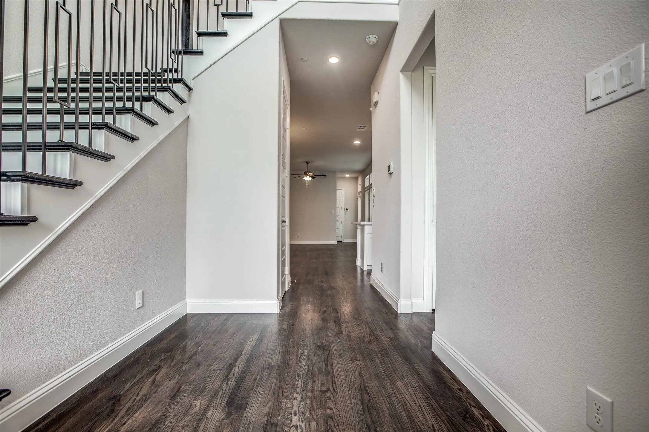 Dark wood-finish flooring with white baseboards, a staircase featuring black metal balusters and dark treads, recessed lighting, and a white interior door