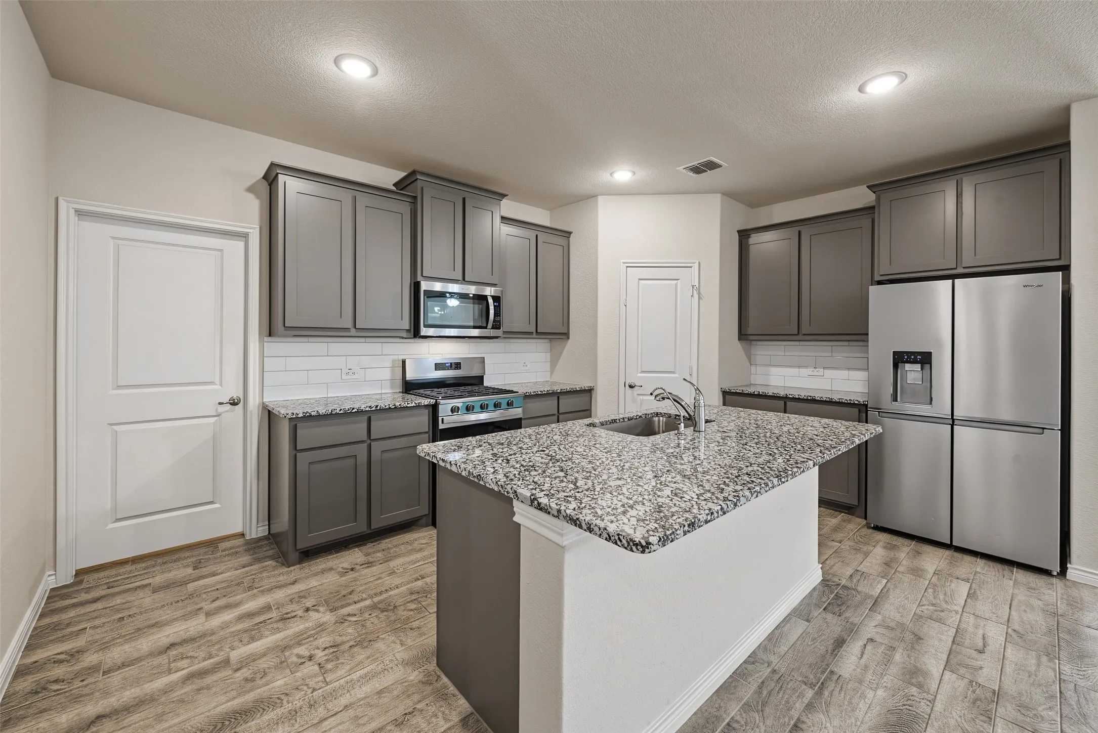 Kitchen featuring an island with a built-in sink, gray cabinetry, stainless steel appliances, porcelain tile flooring and recessed lighting