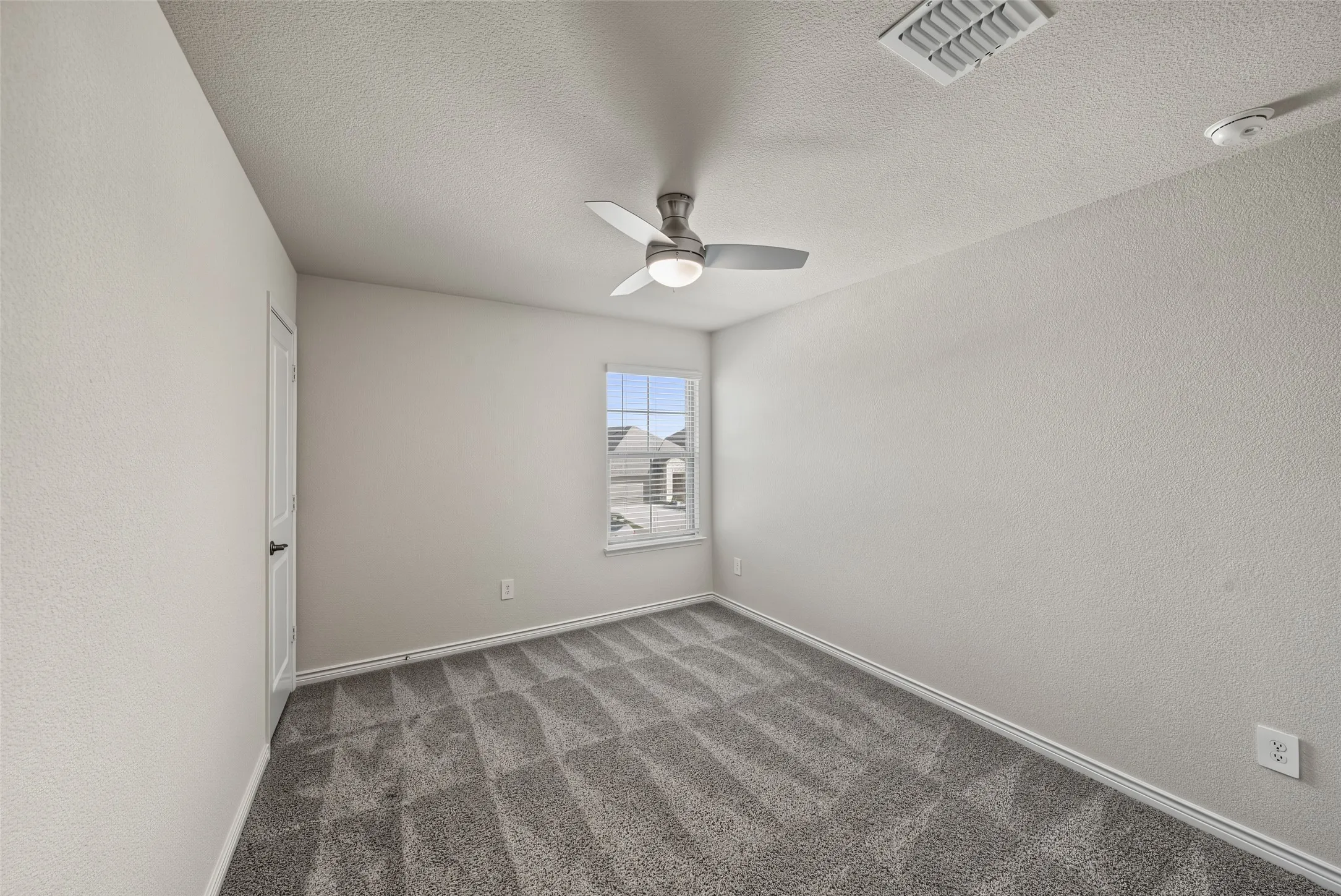 Textured neutral walls, gray carpeting, single window with white trim, white door with dark hardware, and a ceiling fan with integrated lighting