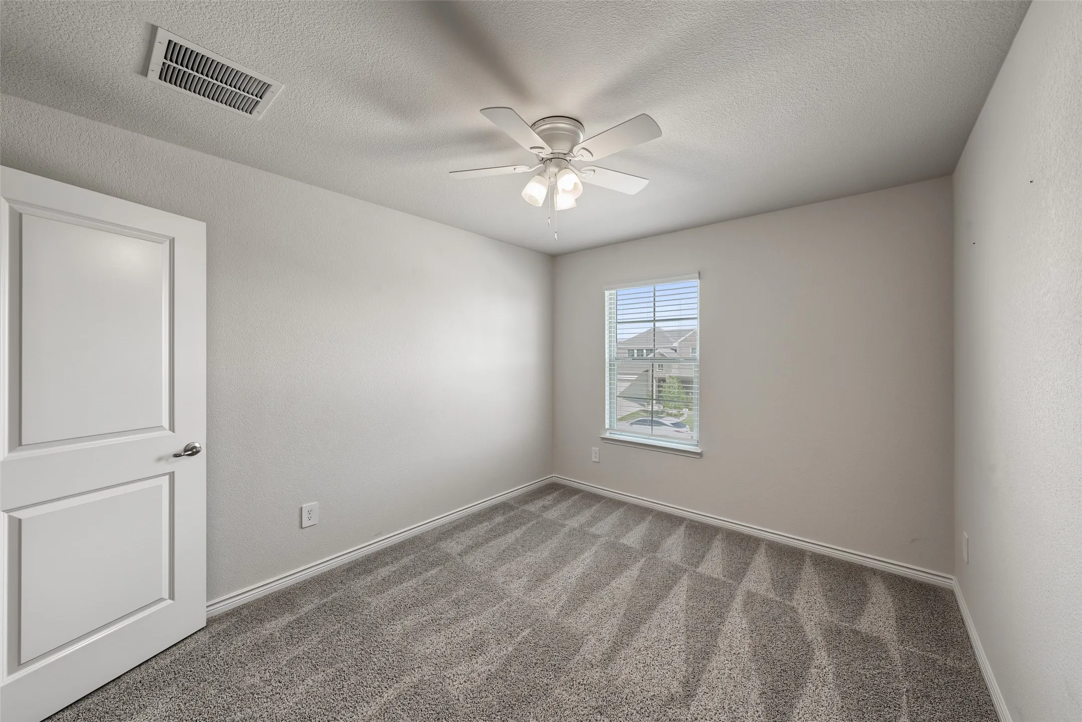 Neutral wall tones, gray patterned carpet, ceiling fan with light fixture, single window with blinds, white paneled door