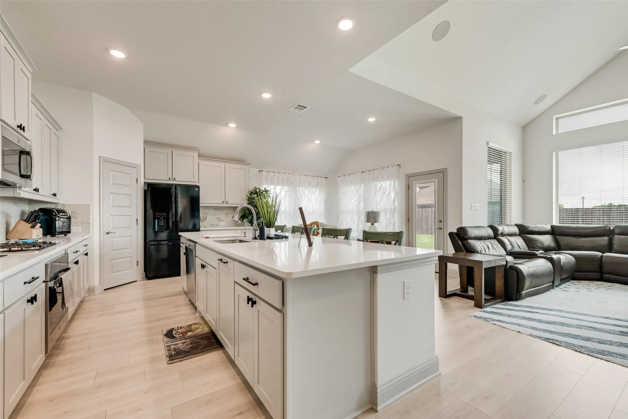 Recessed lighting, wood-finish flooring, white cabinetry with dark hardware, kitchen island with integrated sink, and vaulted ceiling