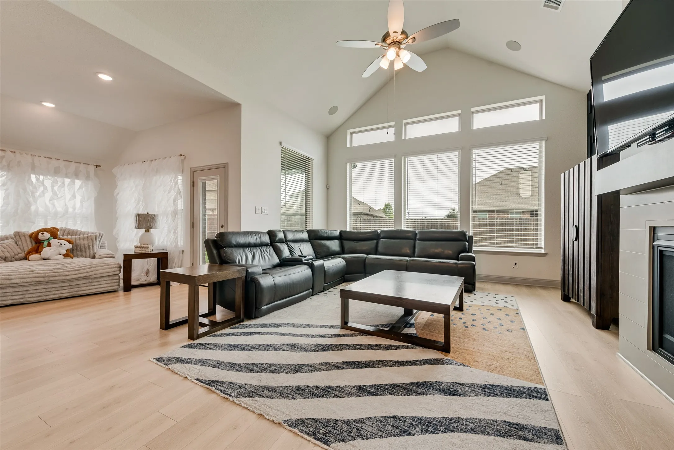 Vaulted ceiling with recessed lighting, light wood-finish flooring, multiple large windows, ceiling fan with integrated lighting, and a built-in fireplace