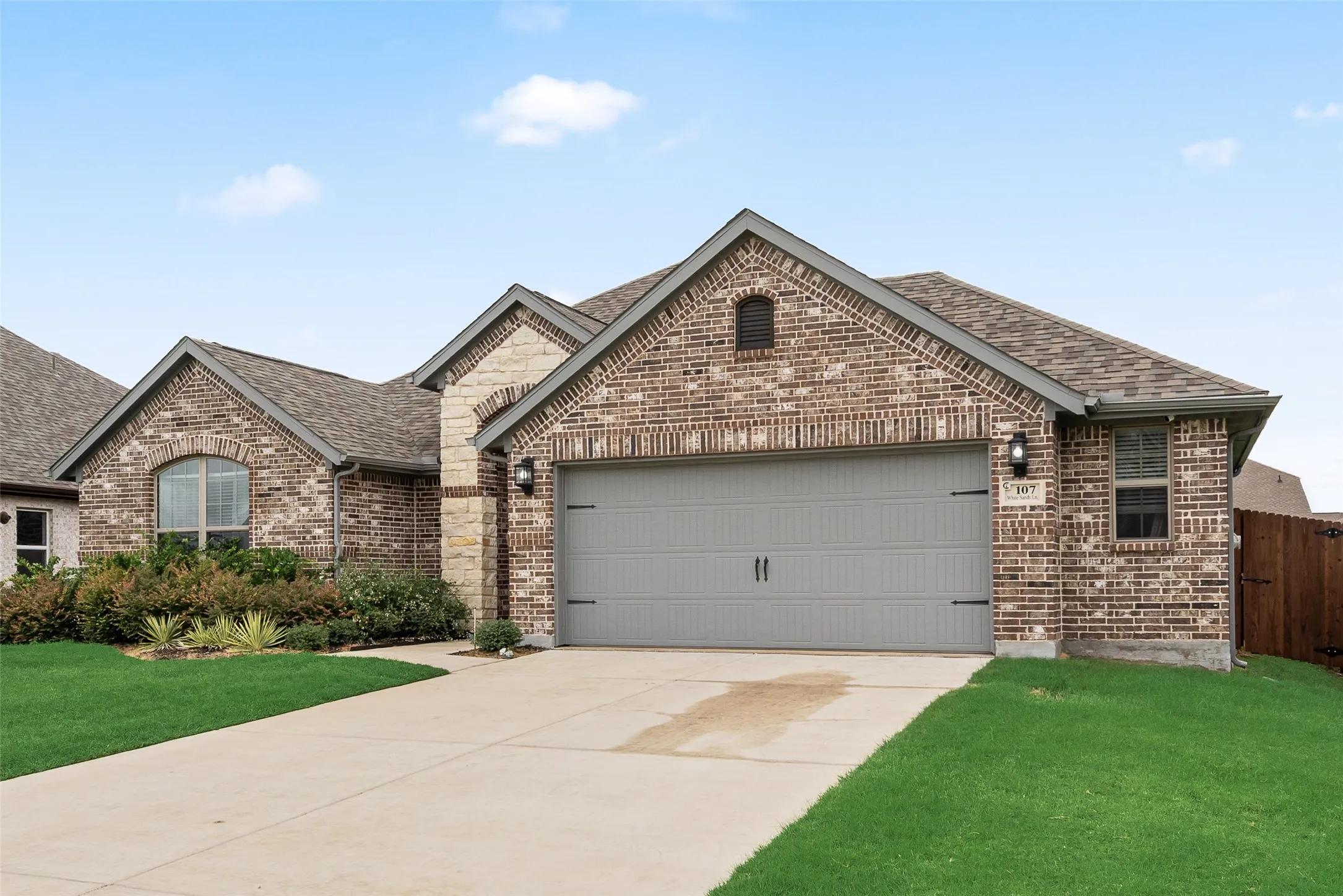 Brick facade, attached garage with a carriage-style door, concrete driveway, and established front landscaping