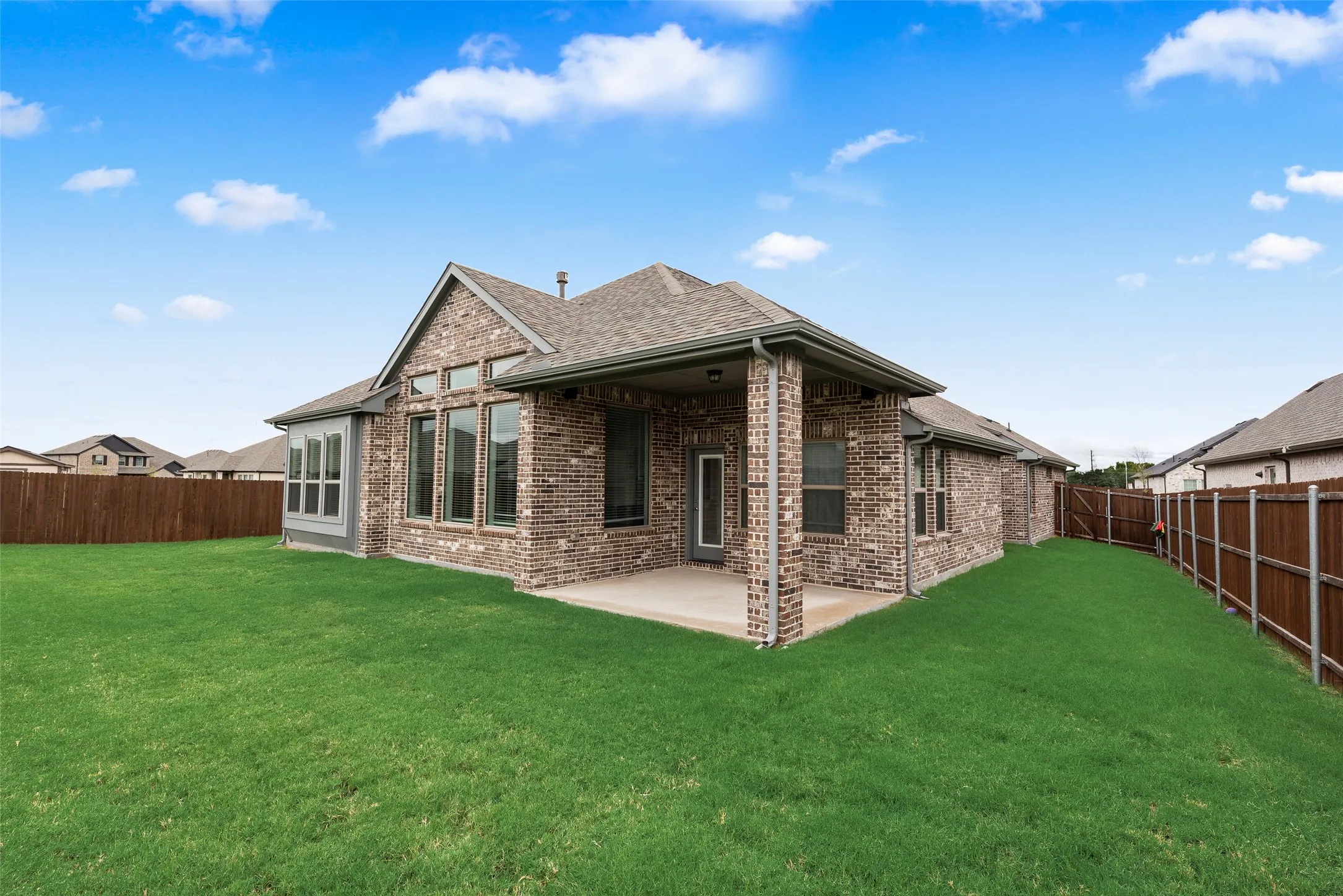 Expansive green lawn with a wood fence, brick exterior, covered patio, and large windows