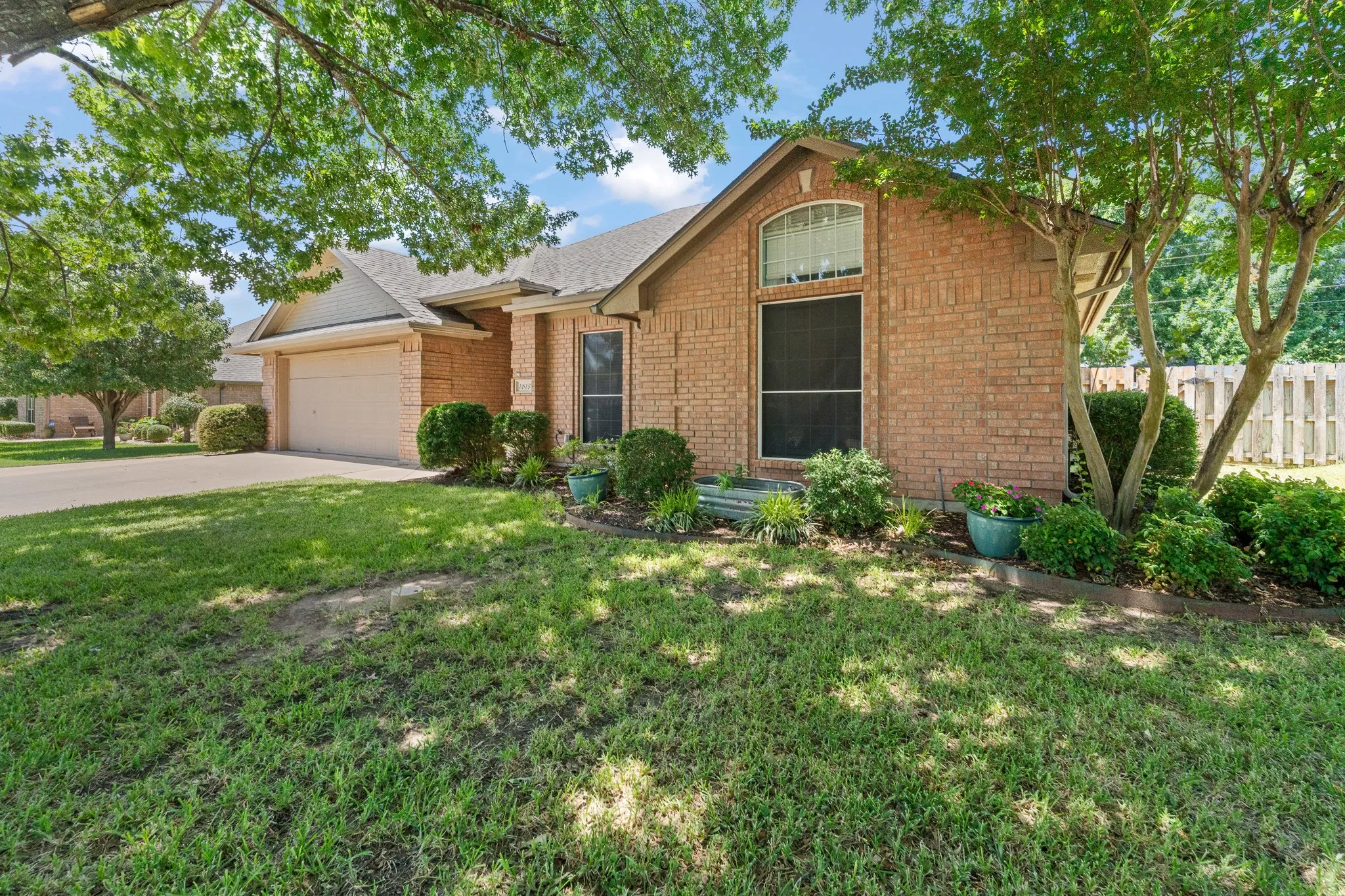 Brick exterior with an attached garage, landscaped front yard, mature trees, and a concrete driveway