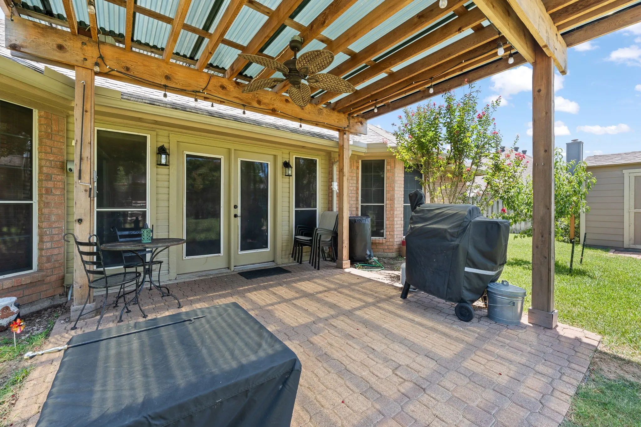 Expansive brick paver patio with a wood pergola and translucent roof, featuring a ceiling fan and string lighting