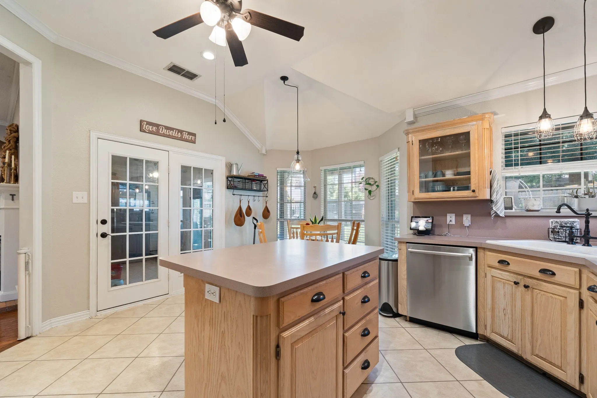 Kitchen featuring an island with built-in storage, light wood cabinetry, stainless steel dishwasher, tile flooring, and a vaulted ceiling with a ceiling fan