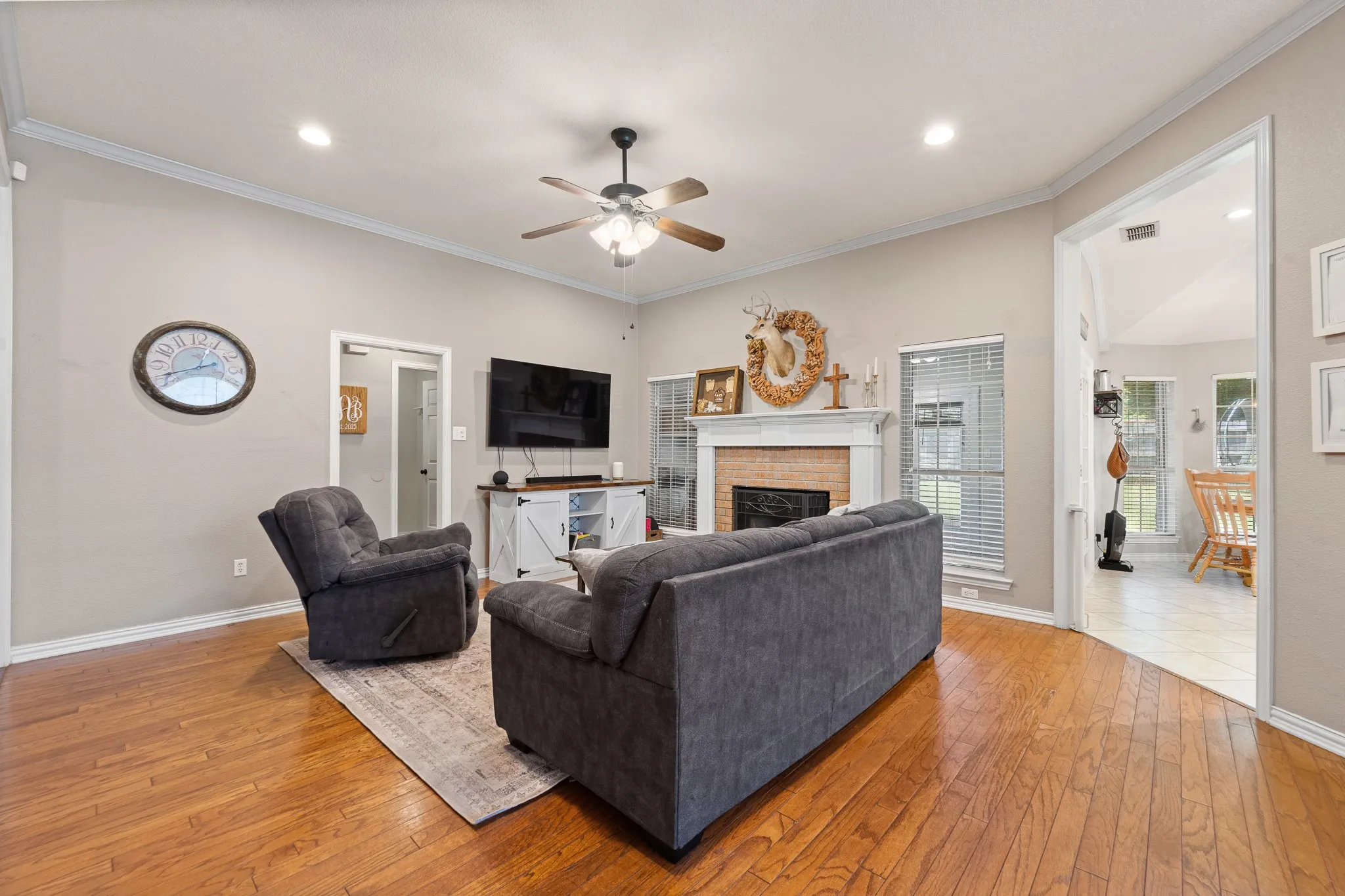 Wood-finish flooring, vaulted ceiling, crown molding, ceiling fan with light fixture, and a brick fireplace with a white mantel