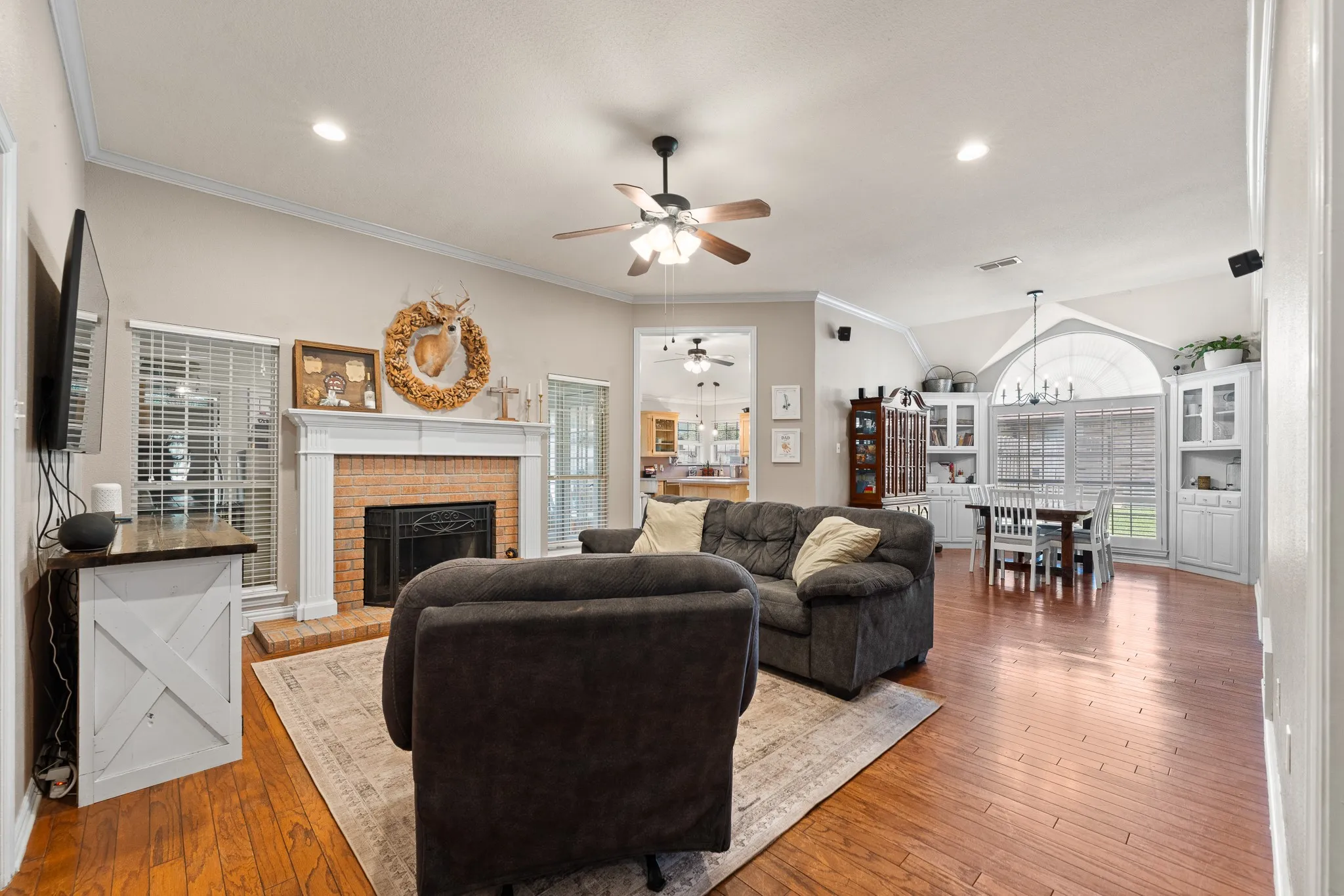 Hardwood flooring throughout, brick fireplace with white mantel, ceiling fan, crown molding, and recessed lighting