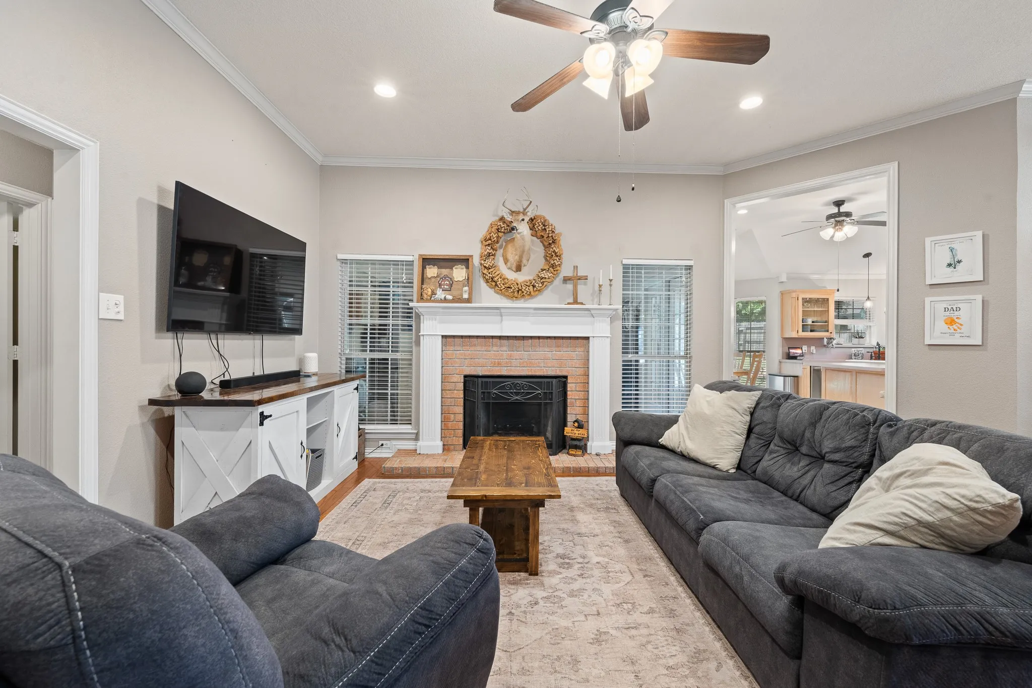 Brick-faced fireplace with a white mantel, ceiling fan with wood-finish blades, recessed lighting, and crown molding