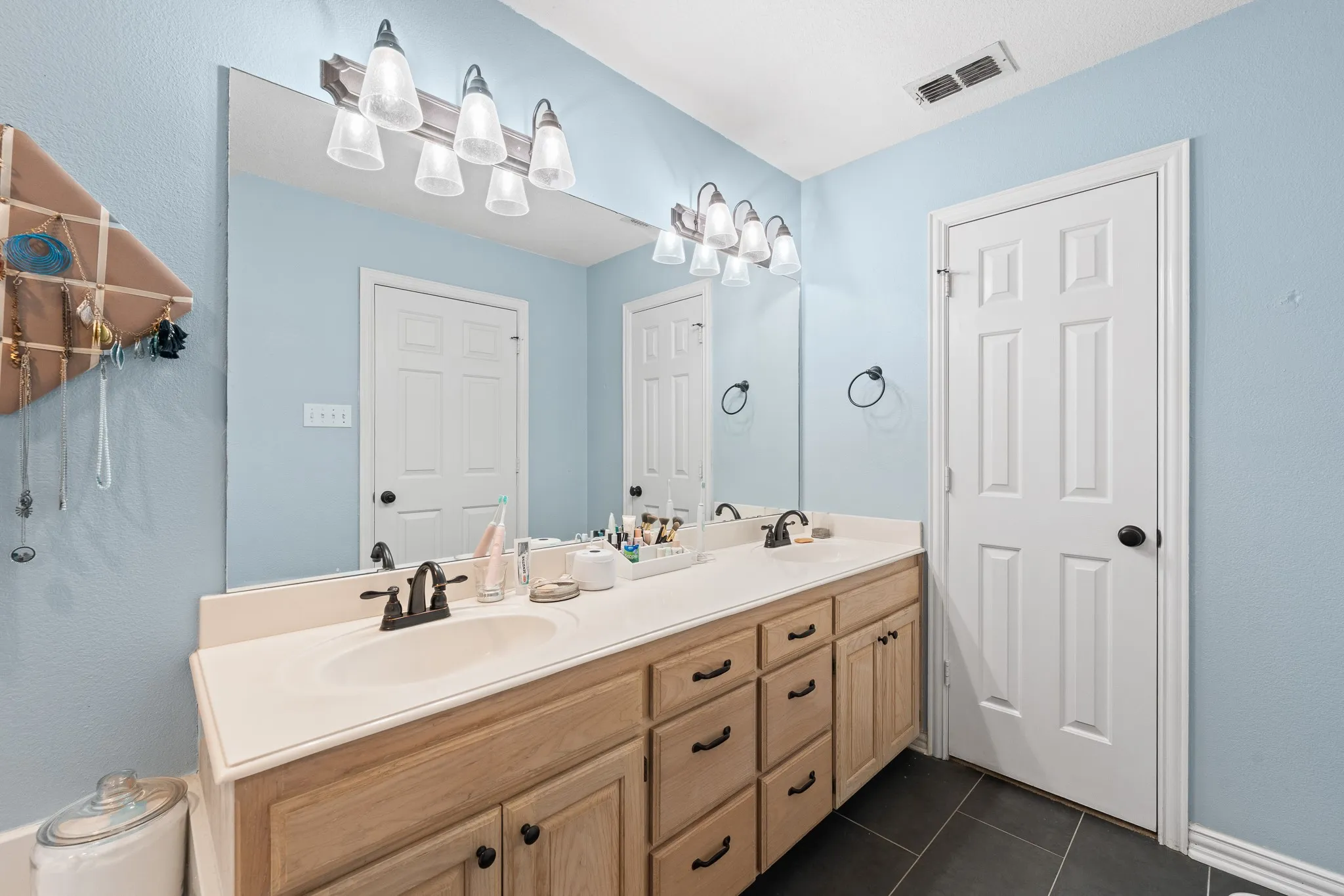 Dual vanity with light wood-finish cabinetry, integrated sinks, dark bronze fixtures, and a full-width mirror