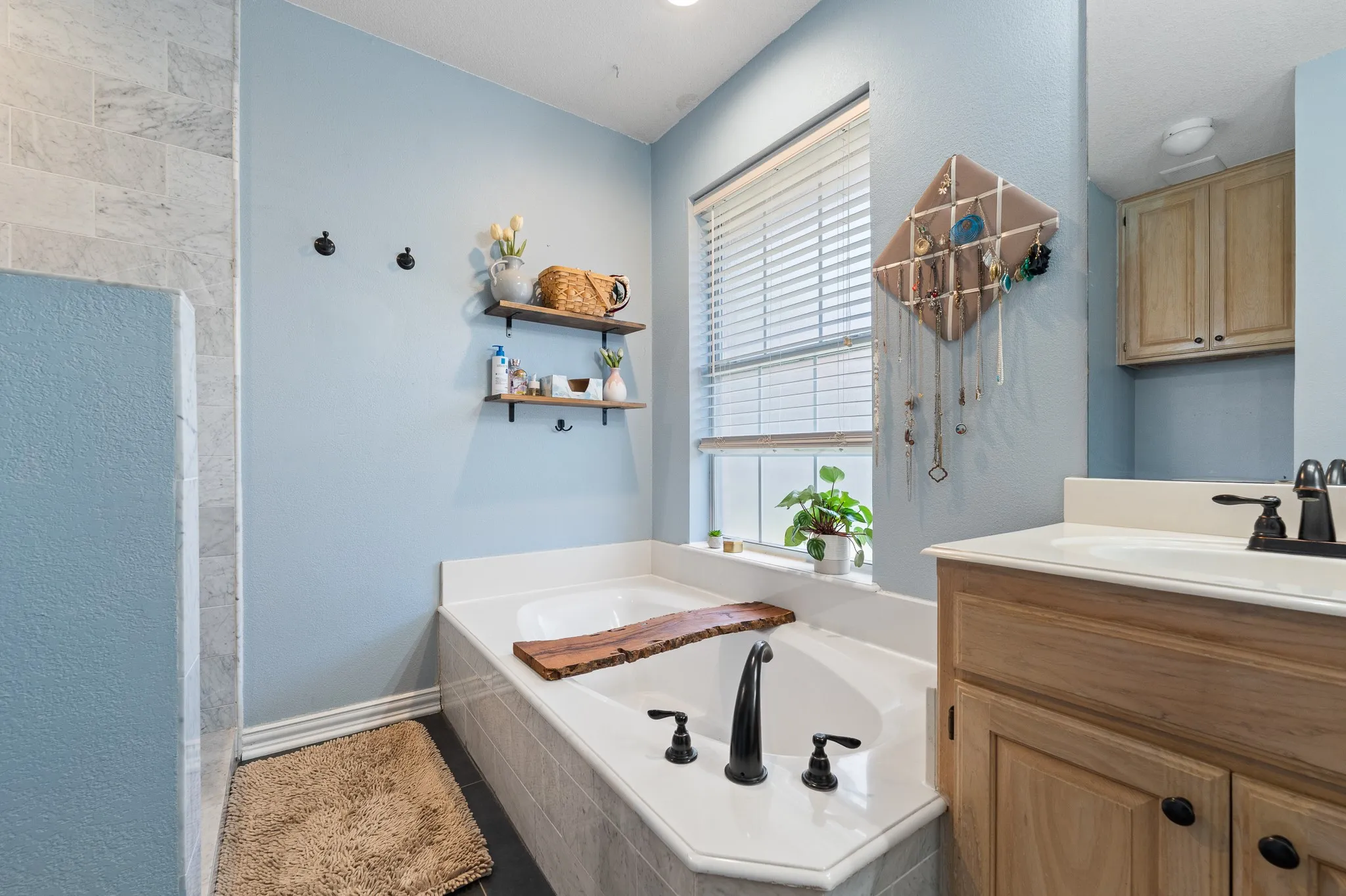 Built-in garden tub with dark bronze fixtures, window with blinds, light wood-finish vanity with countertop, and overhead built-in cabinetry