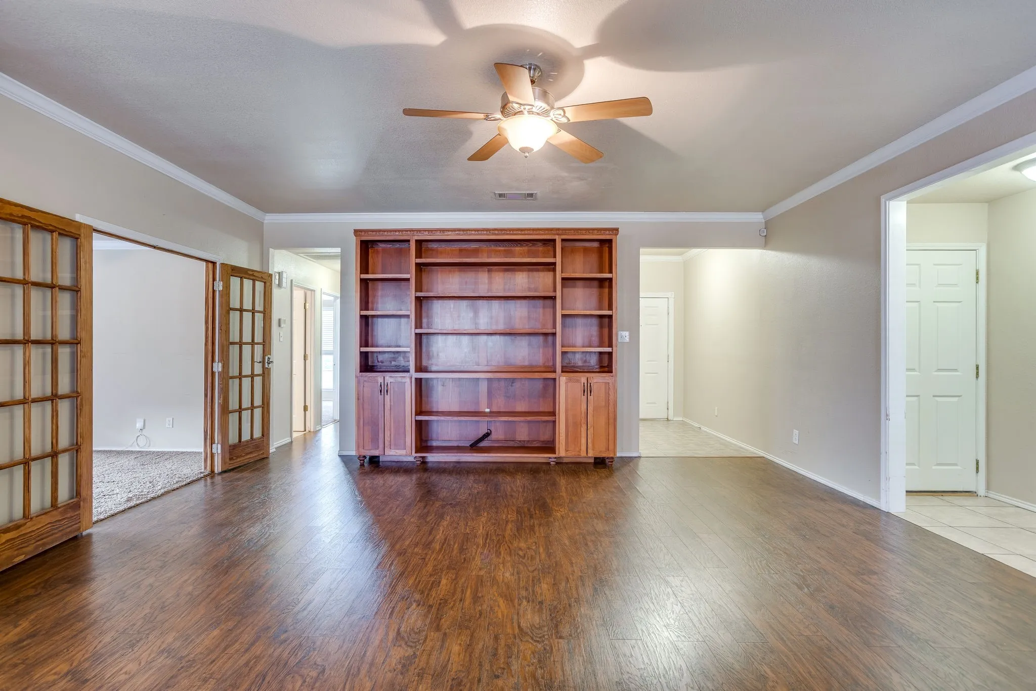 Wood-finish flooring, built-in wooden shelving, double French doors, ceiling fan with integrated lighting, and crown molding