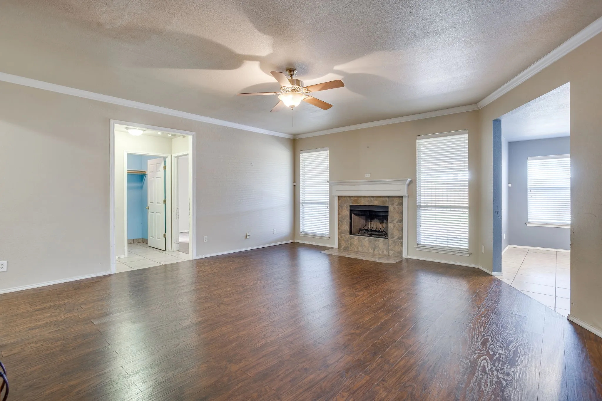 Wood-finish flooring, ceiling fan, fireplace with tile surround, and multiple windows with blinds