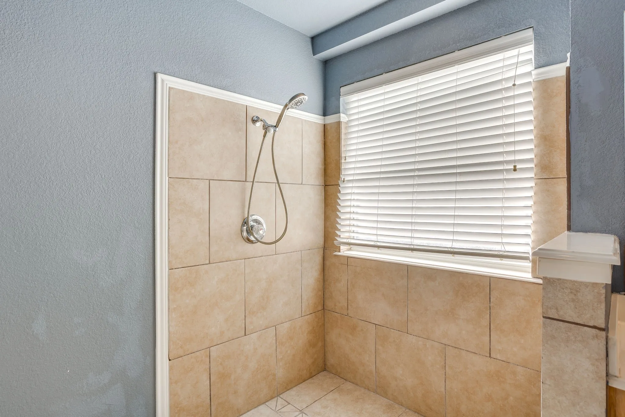 Tiled shower enclosure featuring a handheld showerhead with chrome fixtures, a window with horizontal blinds, and a tiled half-wall