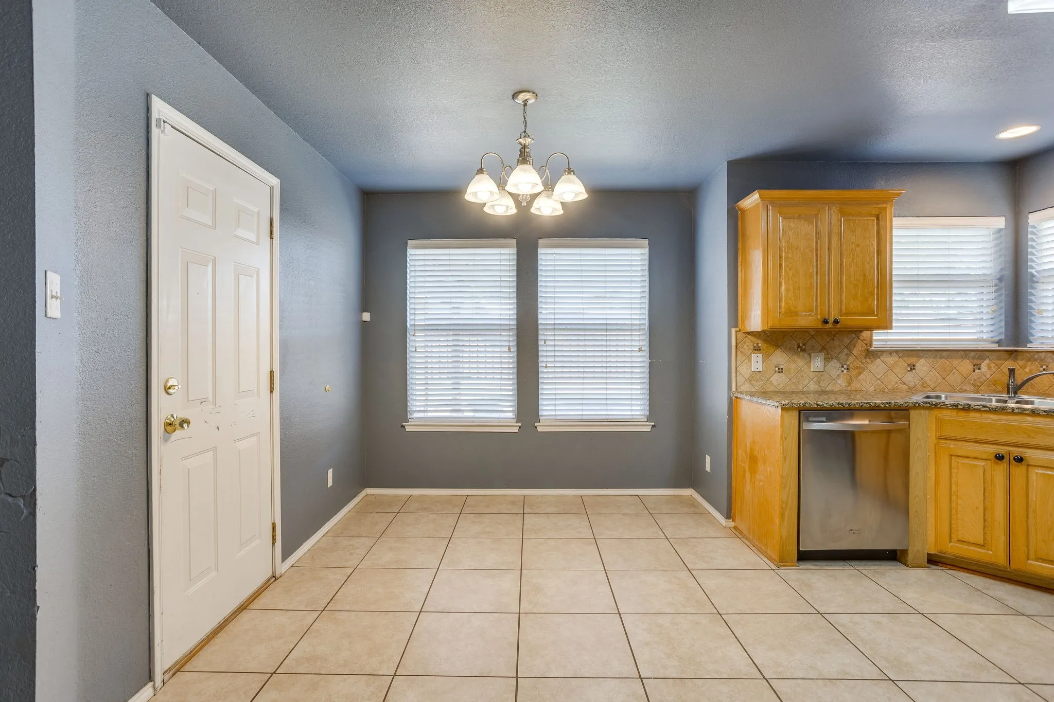 Tiled flooring, warm wood cabinetry, stainless steel dishwasher, decorative tile backsplash, and a ceiling-mounted lighting fixture