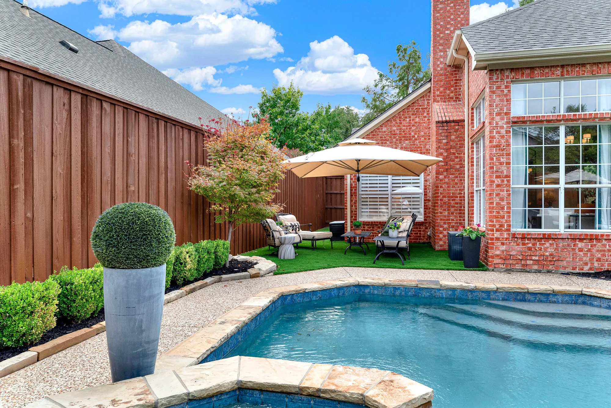Curved pool contours harmoniously balance the surrounding patio areas and dark mulch garden borders. Take a refreshing dip on hot summer days before enjoying dinner alfresco under bright blue skies.