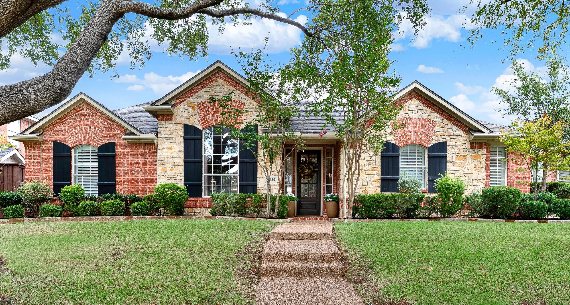 Welcome to 6749 Glenhollow in Frisco.  A rich mix of warm brick and natural stone masonry framing black shuttered windows gives this single-story façade classic curb appeal. Step across the walkway as dappled light filters through mature shade trees.