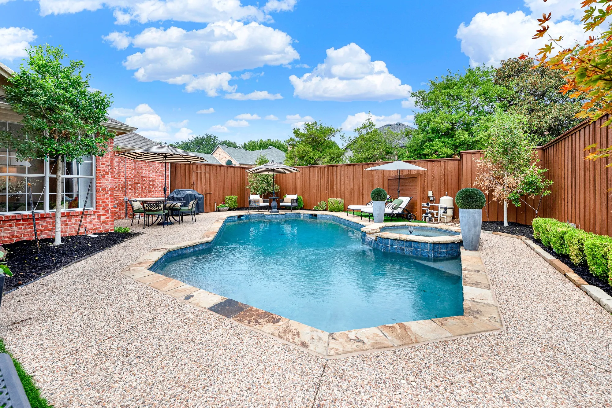 Lush artificial turf in the backyard creates a vibrant, low-maintenance green corner tucked against classic red brick. Savor a peaceful morning coffee beneath shade umbrellas in your serene outdoor sanctuary.