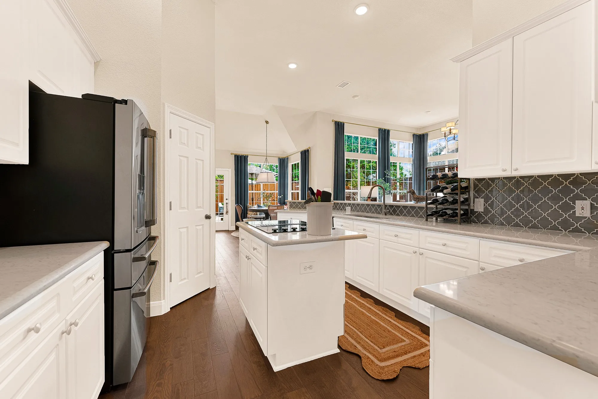 A centered cooktop island enhances functional traffic flow beneath high vaulted ceiling lines. Sip a  quick afternoon espresso while watching sunlight stream through the adjoining living spaces.