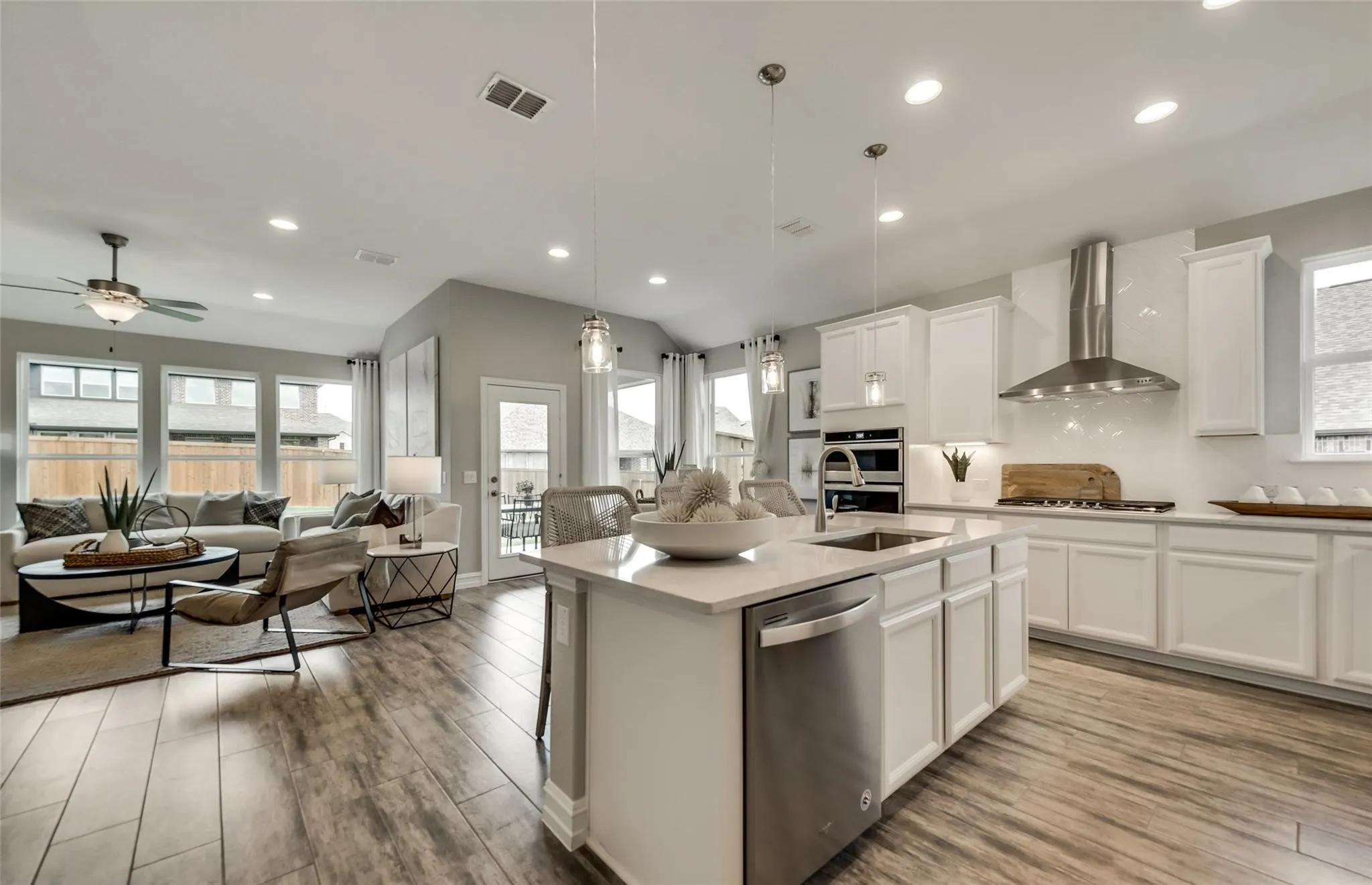 Open-concept design featuring wood-finish flooring, a kitchen island with sink and integrated dishwasher, white shaker cabinetry, and a stainless steel range hood