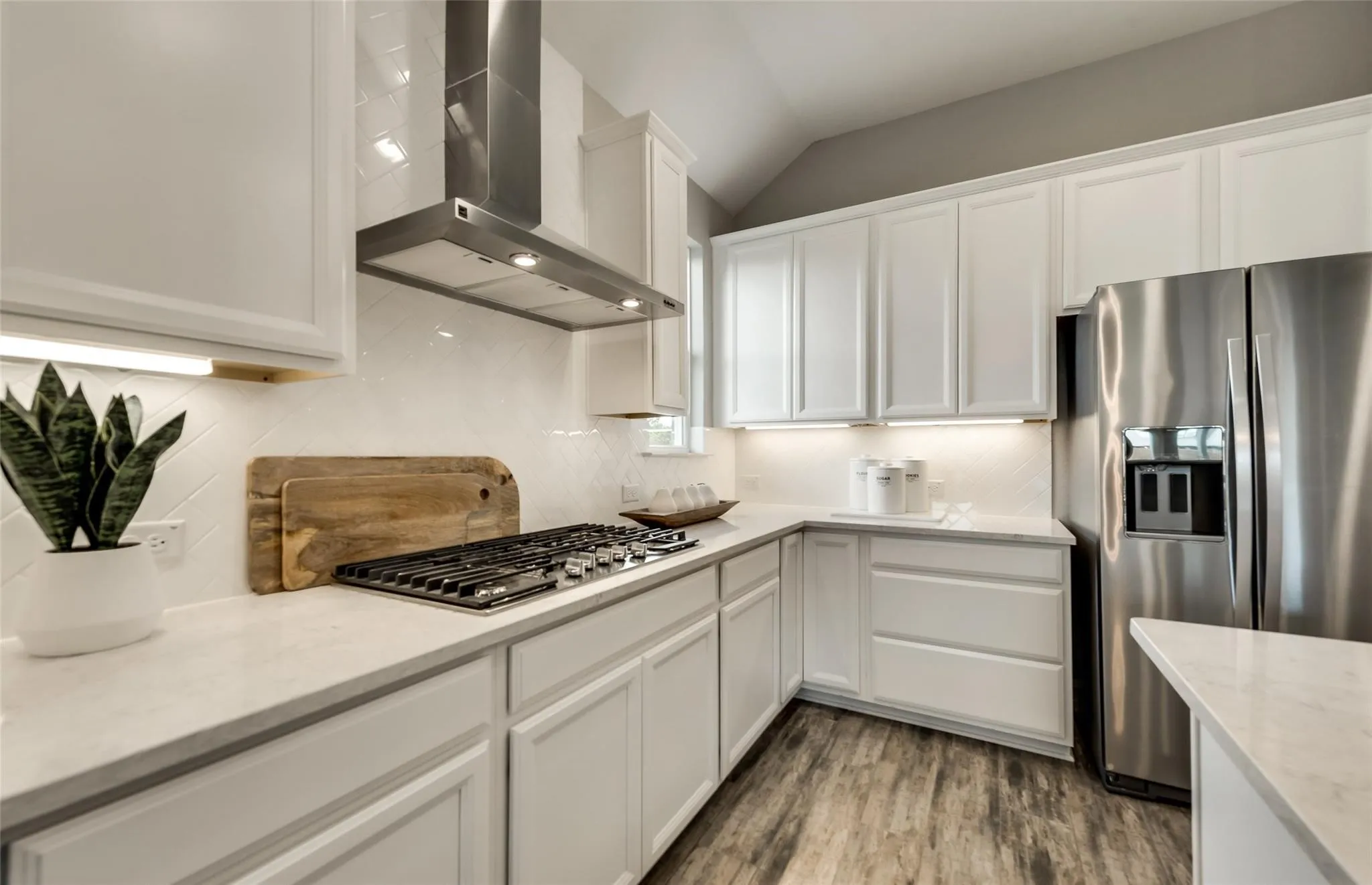 White shaker-style cabinetry with under-cabinet lighting, stainless steel appliances, a built-in gas range with a stainless steel range hood, stone countertops, and wood-finish flooring