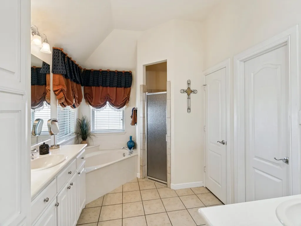 Dual vanity with integrated sinks and white cabinetry, a built-in corner soaking tub, a tiled shower with a glass door, and neutral-toned floor tiling