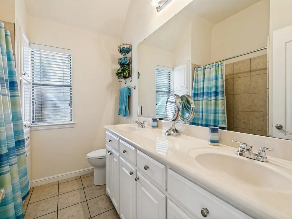 Dual sink vanity with white cabinetry and chrome hardware, a large mirror, a separate toilet area, and tiled flooring