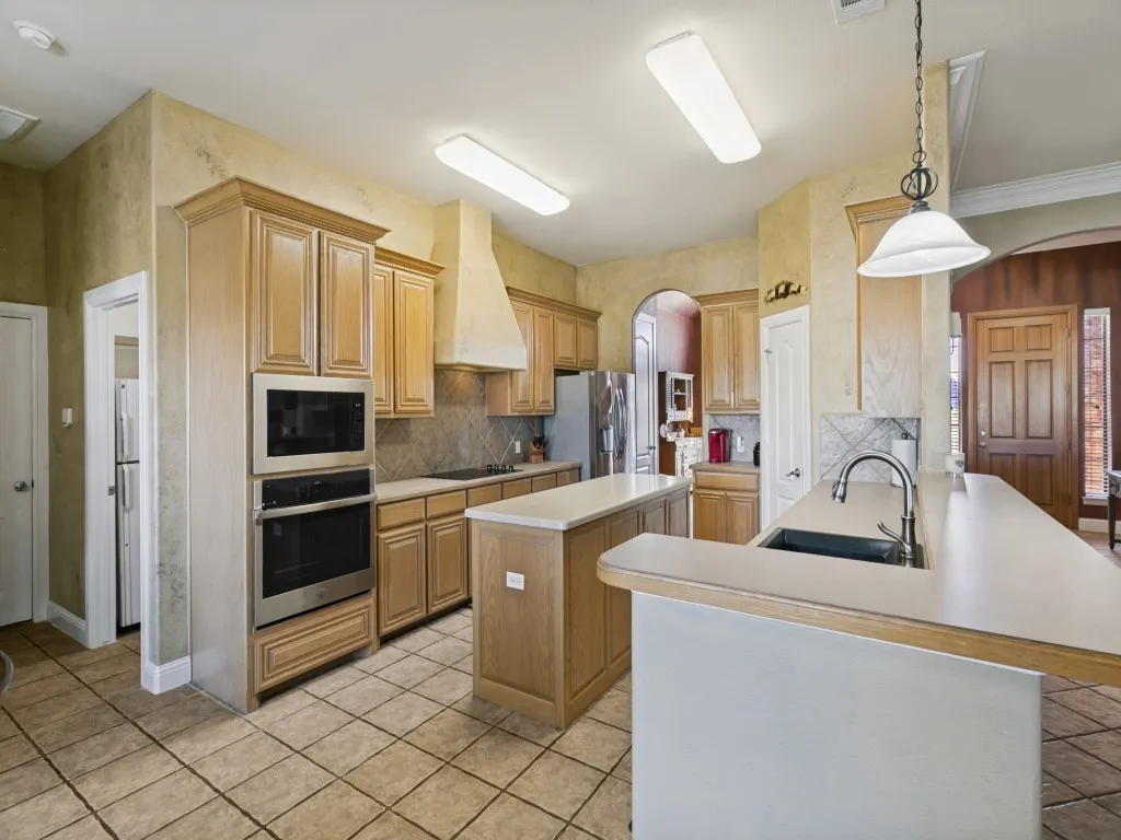 Kitchen with light wood cabinetry, a central island, tile flooring, stainless steel built-in appliances, and a double basin sink