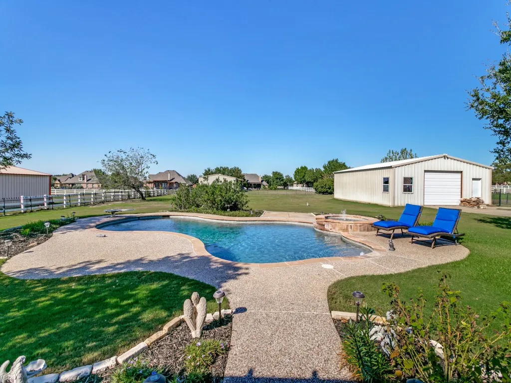 Freeform swimming pool with an integrated spa, expansive concrete pool deck, and a detached metal building with a garage door