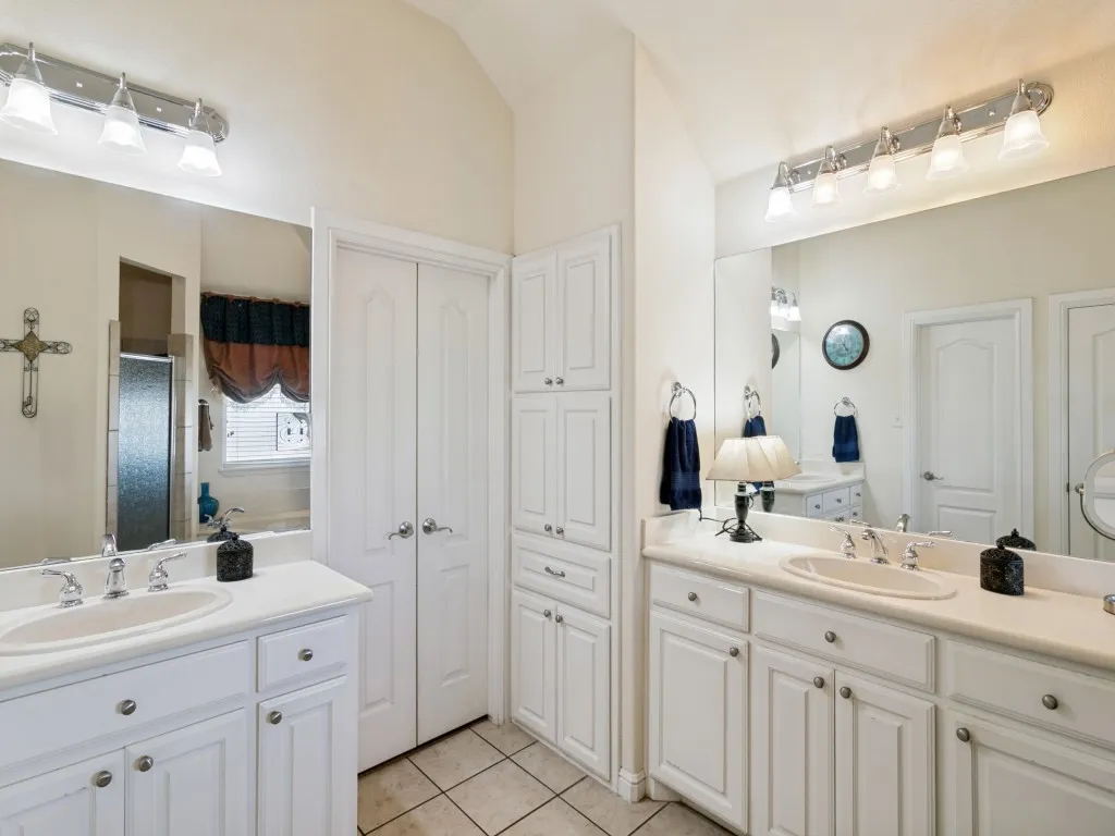 Dual vanities with integrated sinks, extensive white cabinetry, large mirrors, built-in storage tower, and tiled flooring