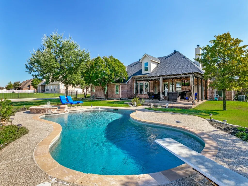 Curved swimming pool with a diving board, integrated hot tub, and a covered patio featuring a wood beam roof