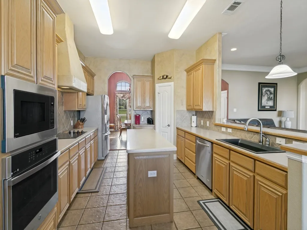 Galley-style kitchen featuring wood cabinetry, tile flooring, stainless steel appliances including double wall ovens, a center island with wood-finish base, and granite countertops with undermount sink and chrome faucet
