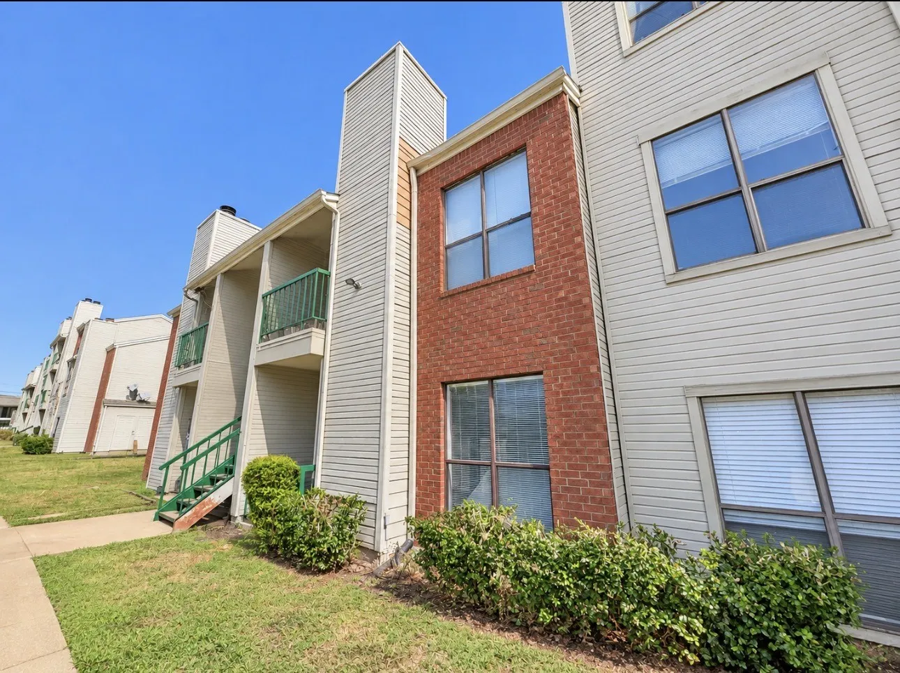 Multilevel building exterior featuring light-colored siding, brick accents, multiple windows with blinds, and balconies with green railings