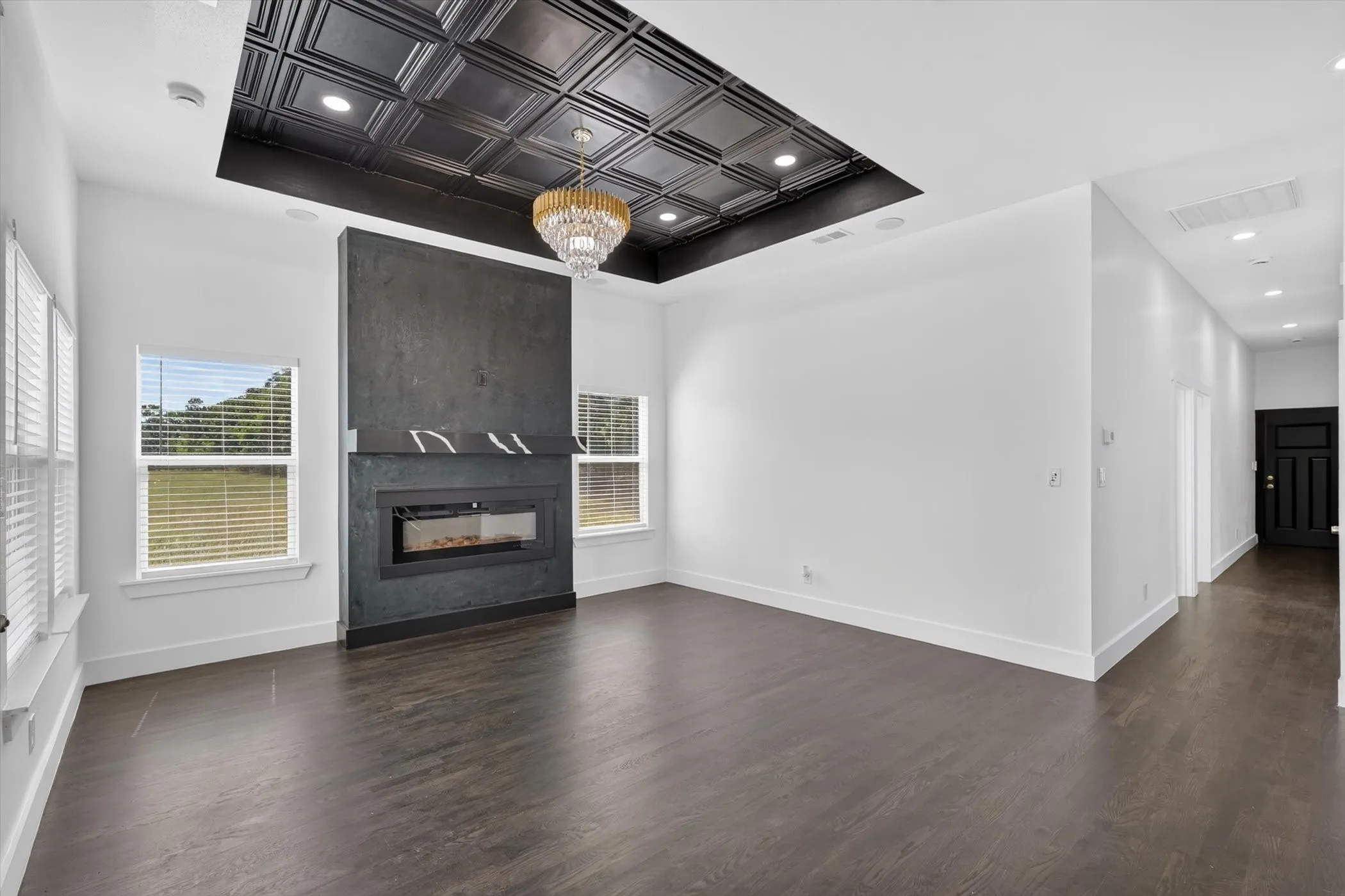 Recessed ceiling featuring a decorative chandelier, electric fireplace with dark-toned surround, two bright windows, and dark wood-finish flooring