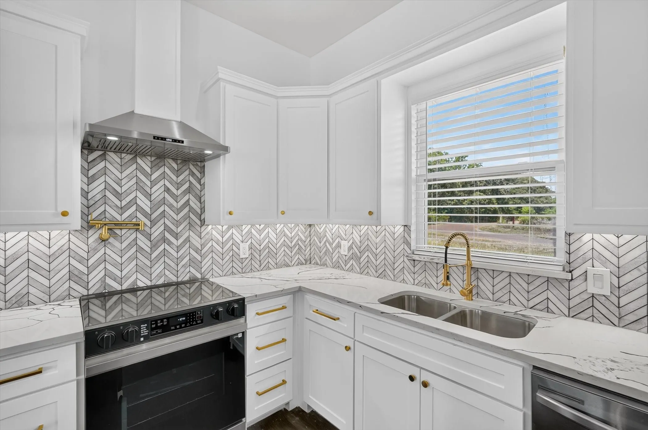 White shaker cabinetry with gold-tone hardware, chevron-patterned tile backsplash, stainless steel appliances, and a double basin sink with a gold-tone gooseneck faucet