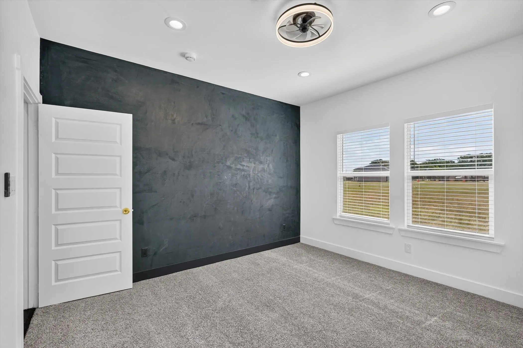 Textured accent wall, recessed lighting, ceiling fan with integrated light, two windows with blinds, and carpeted flooring