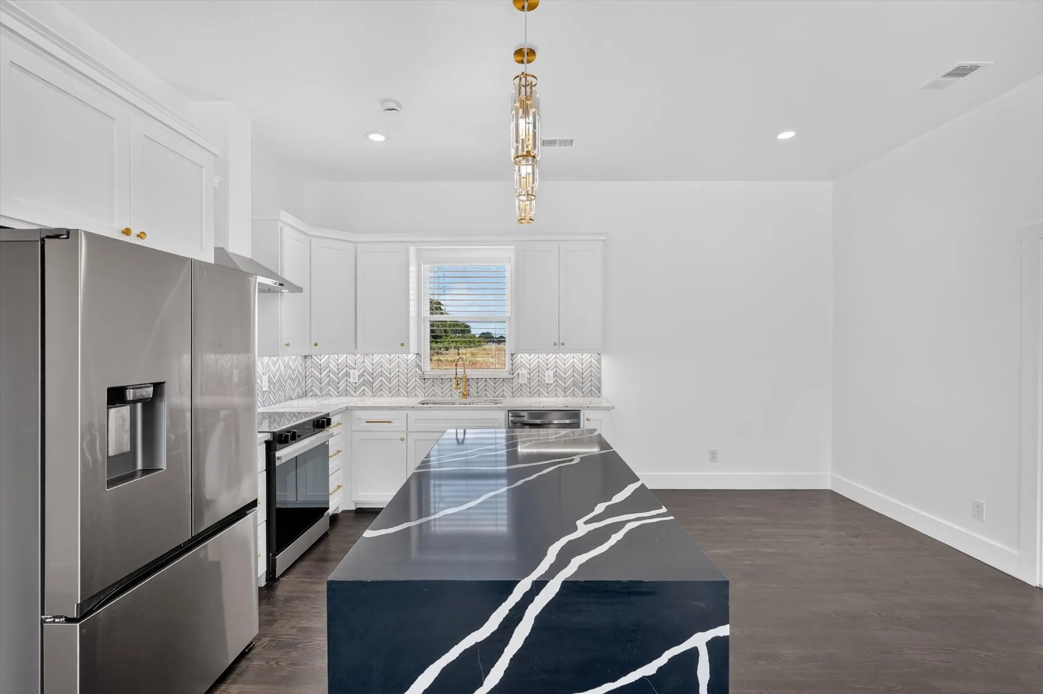 Kitchen featuring a large central island with a dramatic veined countertop, stainless steel appliances, white cabinetry with gold-finish hardware, and a herringbone tile backsplash