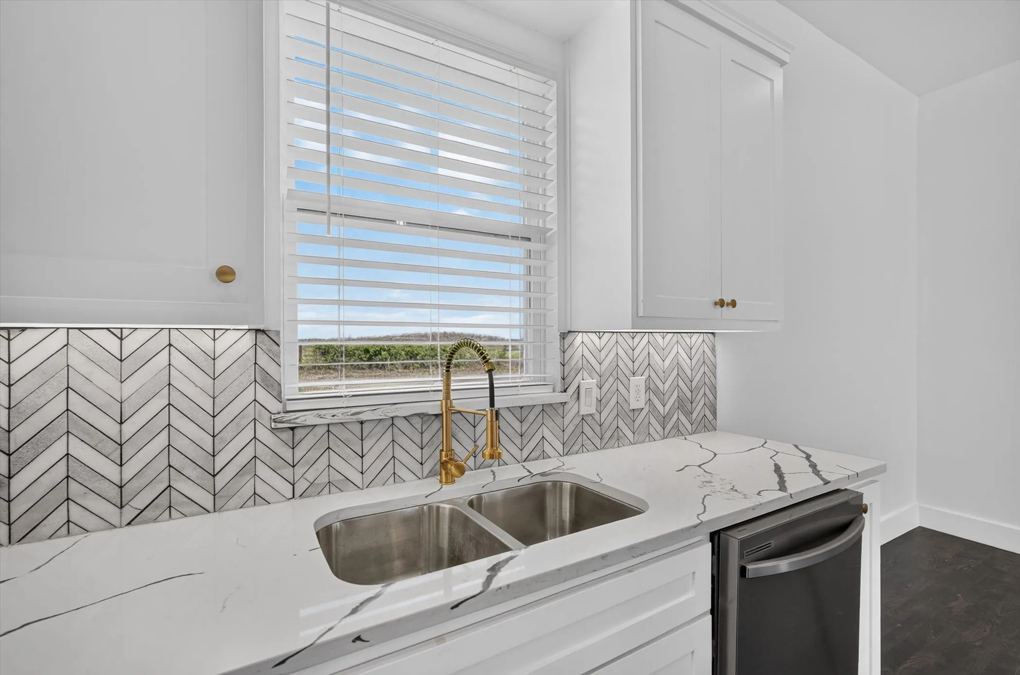 White cabinetry with gold-finish hardware, herringbone tile backsplash, stainless steel double basin sink, gold-finish gooseneck faucet, and white countertops with dark veining