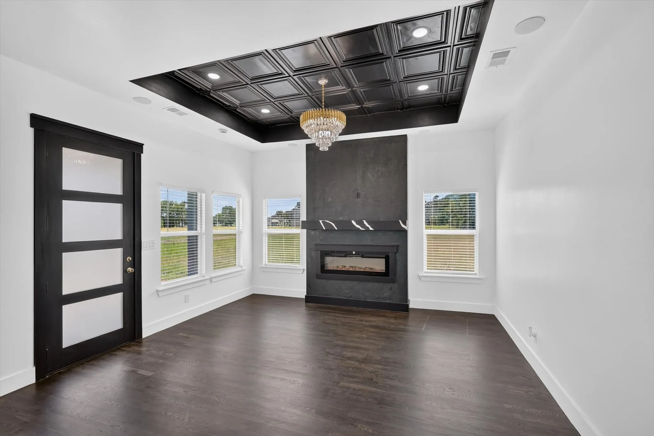 Recessed ceiling with decorative black tile inlay, central chandelier, dark wood-finish flooring, and a built-in fireplace with a stone mantel