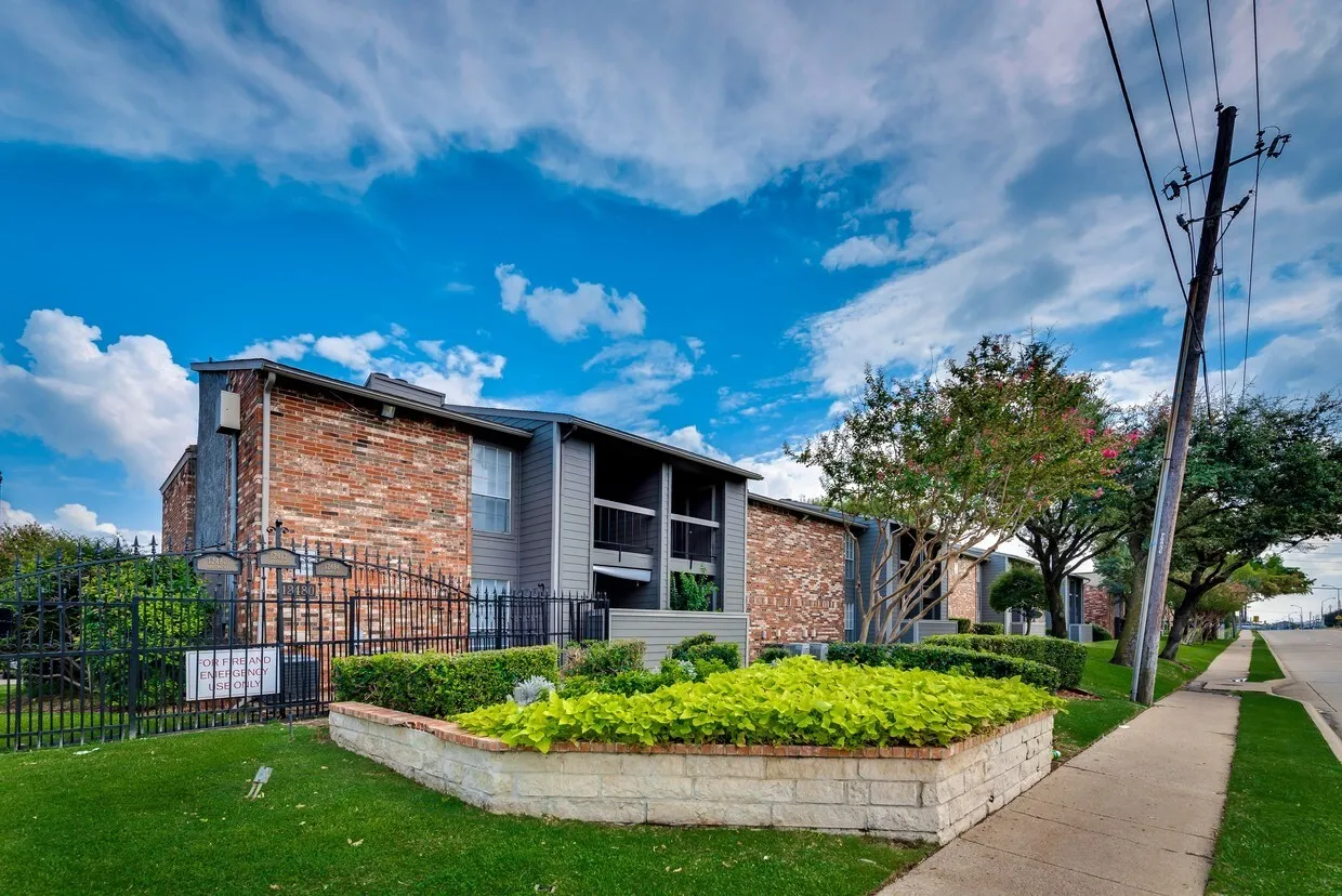 Brick and siding exterior with balconies, featuring a black wrought iron fence, a stone-faced planter with vibrant foliage, and mature trees lining the property