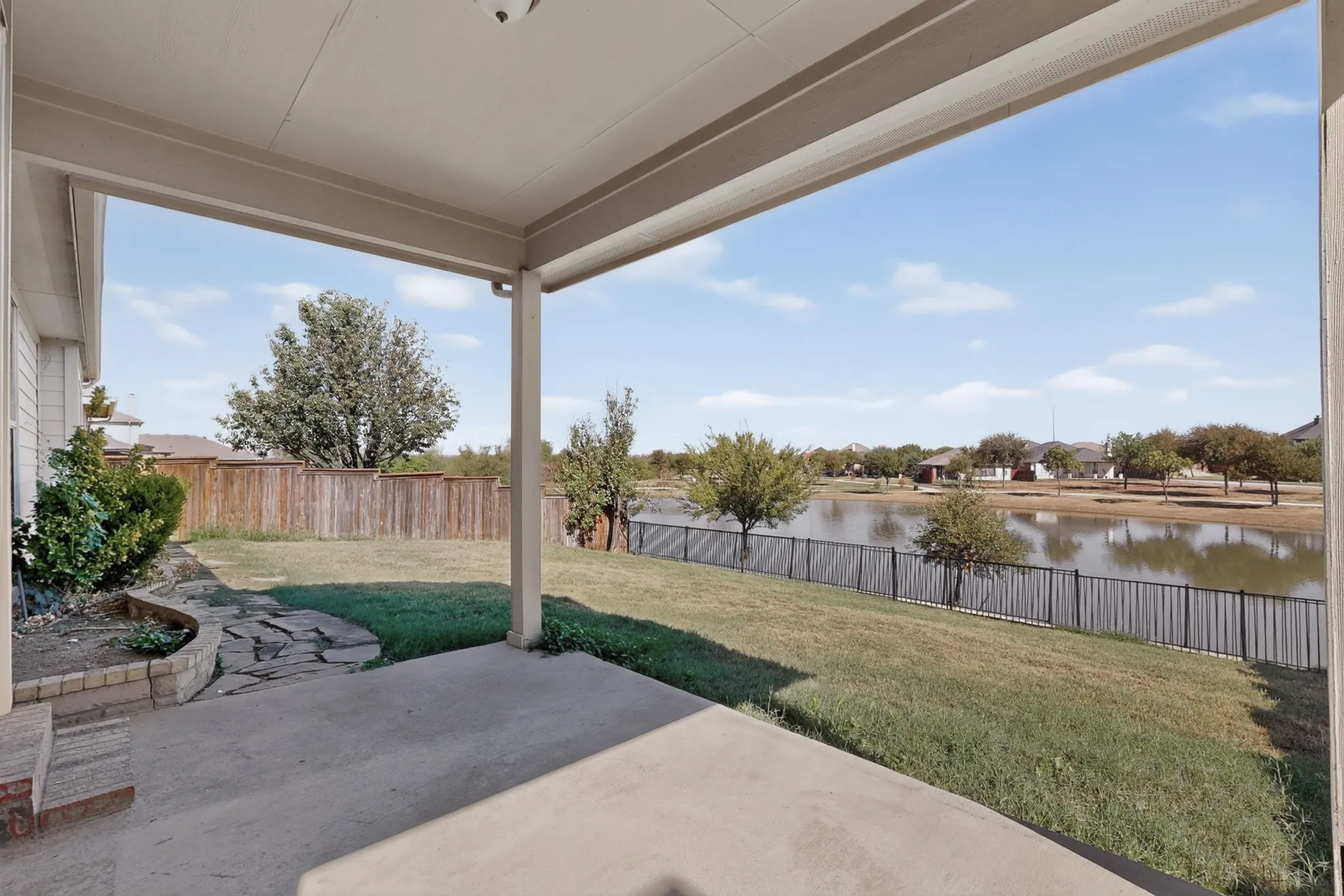 Covered patio with concrete flooring, stone-lined landscaping, and a fenced backyard overlooking a body of water