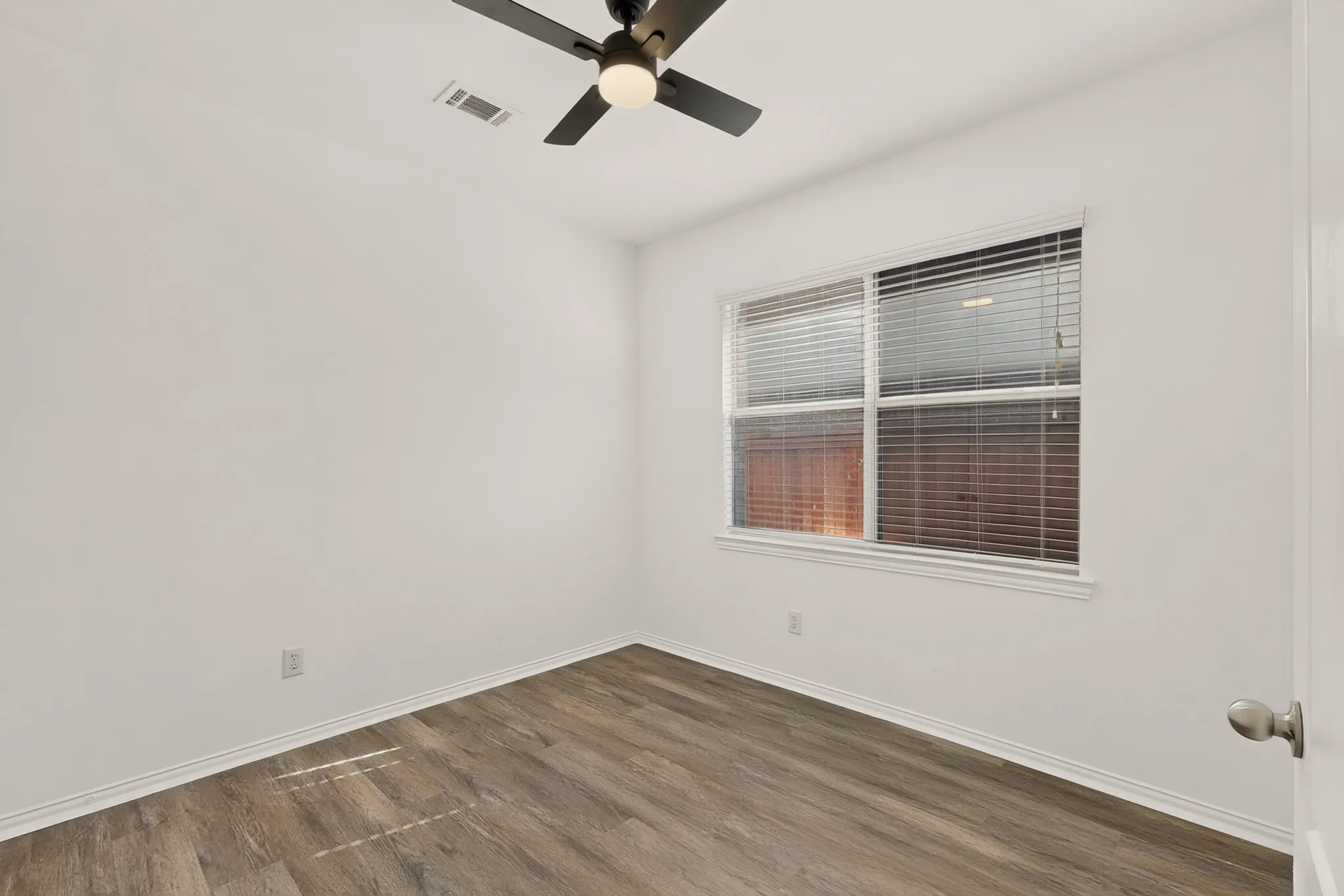 Wood-finish flooring, white walls, double window with blinds, ceiling fan with integrated lighting, and a white door with a satin nickel knob