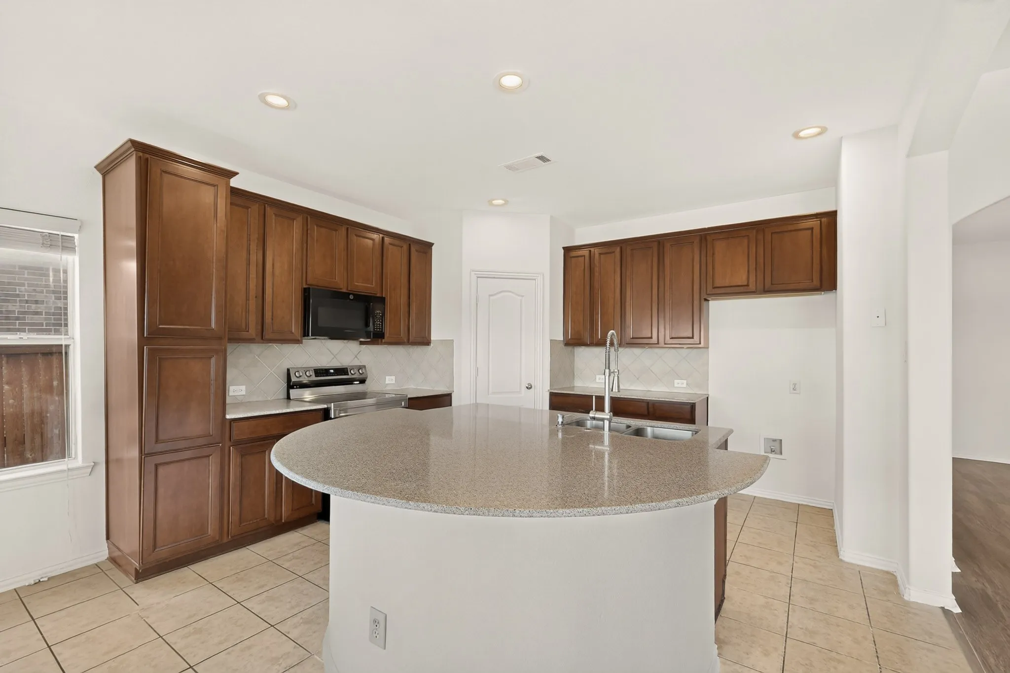 Recessed lighting illuminates a kitchen featuring a curved island with a double basin sink, wood-finish cabinetry, and stainless steel appliances
