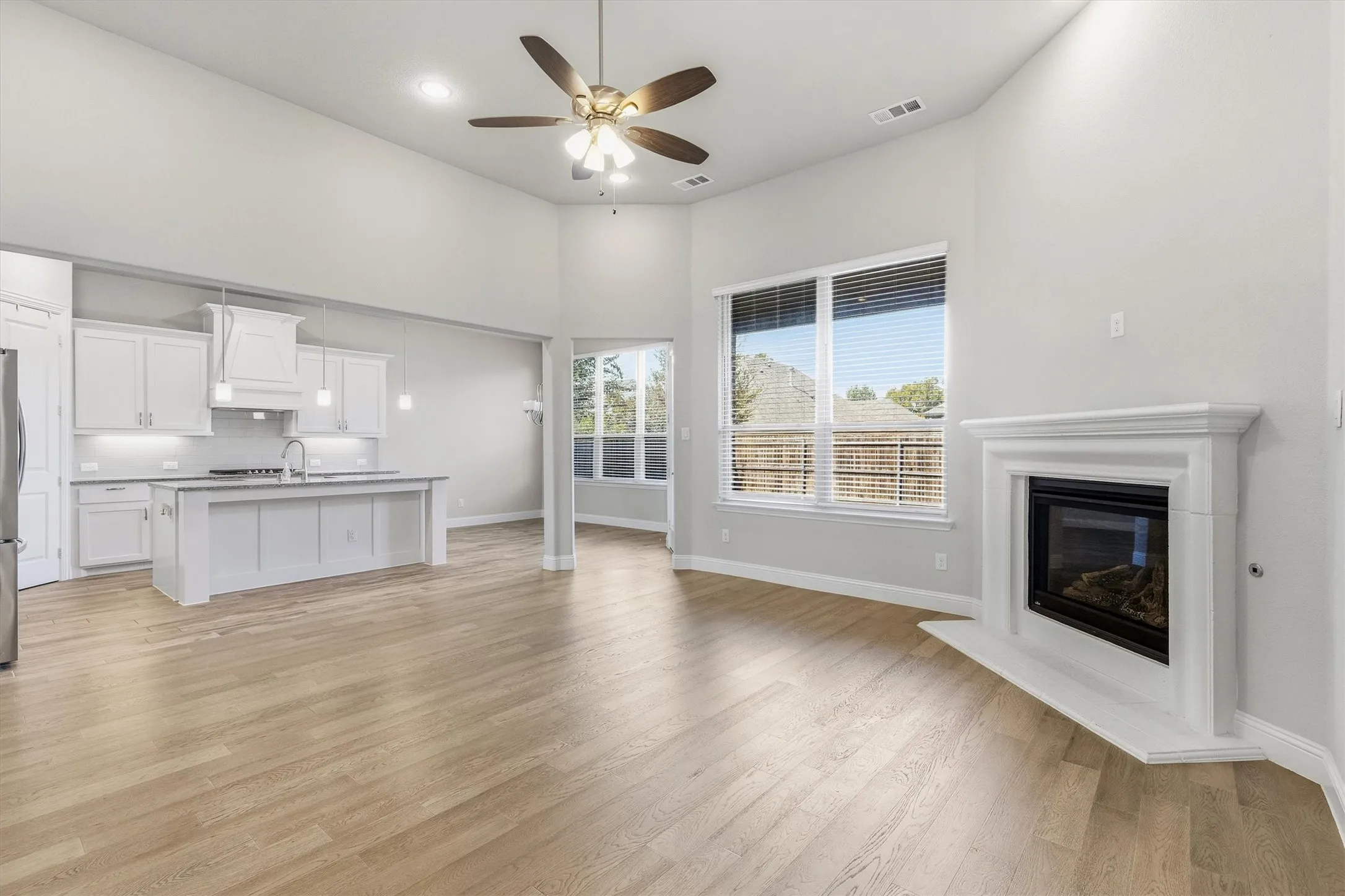 Open-concept living space featuring wood-finish flooring, a white cabinetry kitchen with stainless steel appliances, a fireplace with white surround, ceiling fan with integrated lighting, multiple large windows with views of exterior landscapes, and glass sliding doors providing access to an outdoor deck