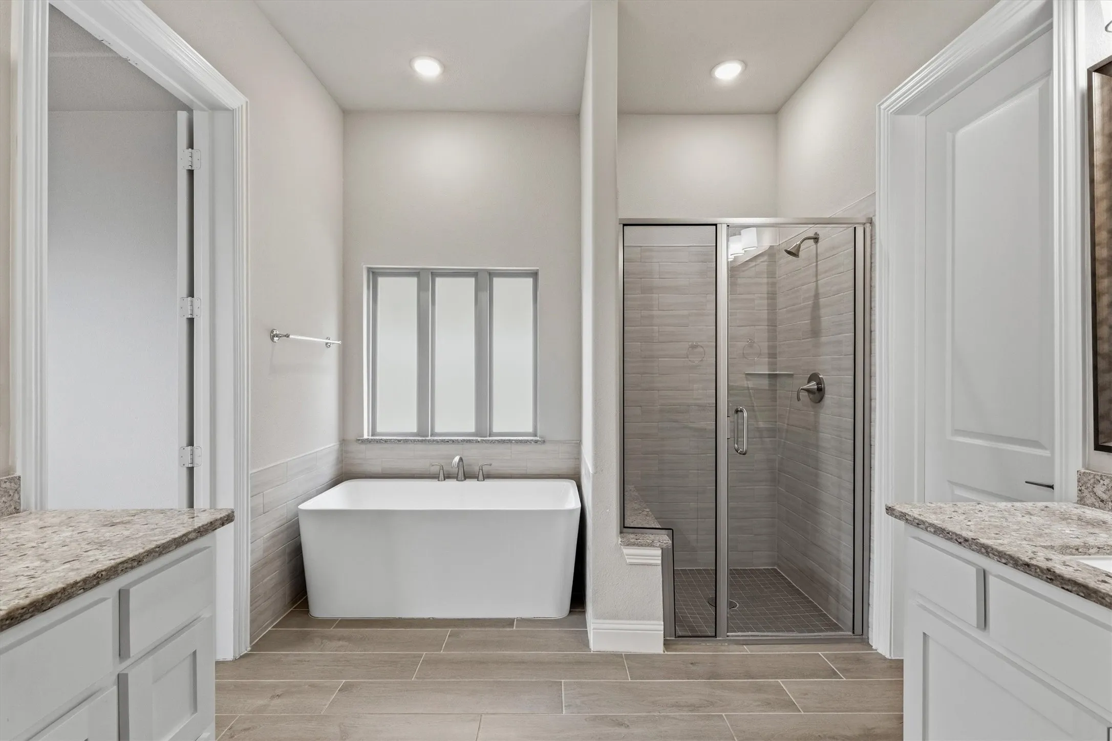 Freestanding bathtub with a tiled surround, separate glass-enclosed shower with a built-in bench, dual vanities with stone countertops, and wood-finish flooring