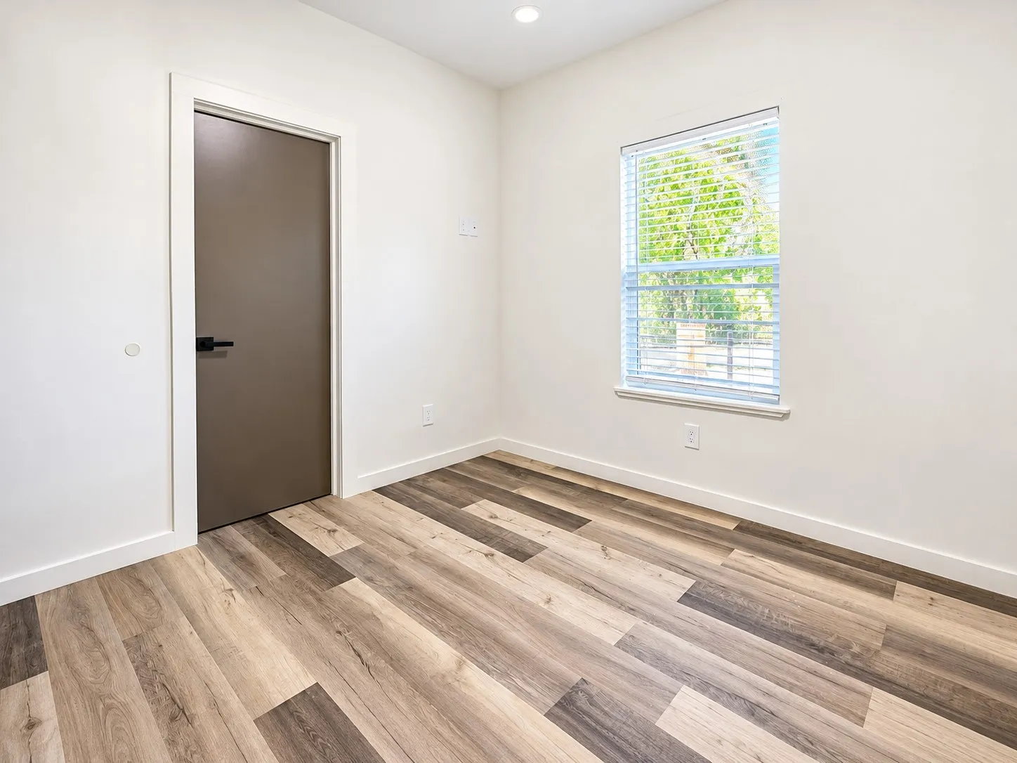 Wood-finish flooring, recessed ceiling lighting, a window with blinds, a modern door with dark hardware, and white wall finishes